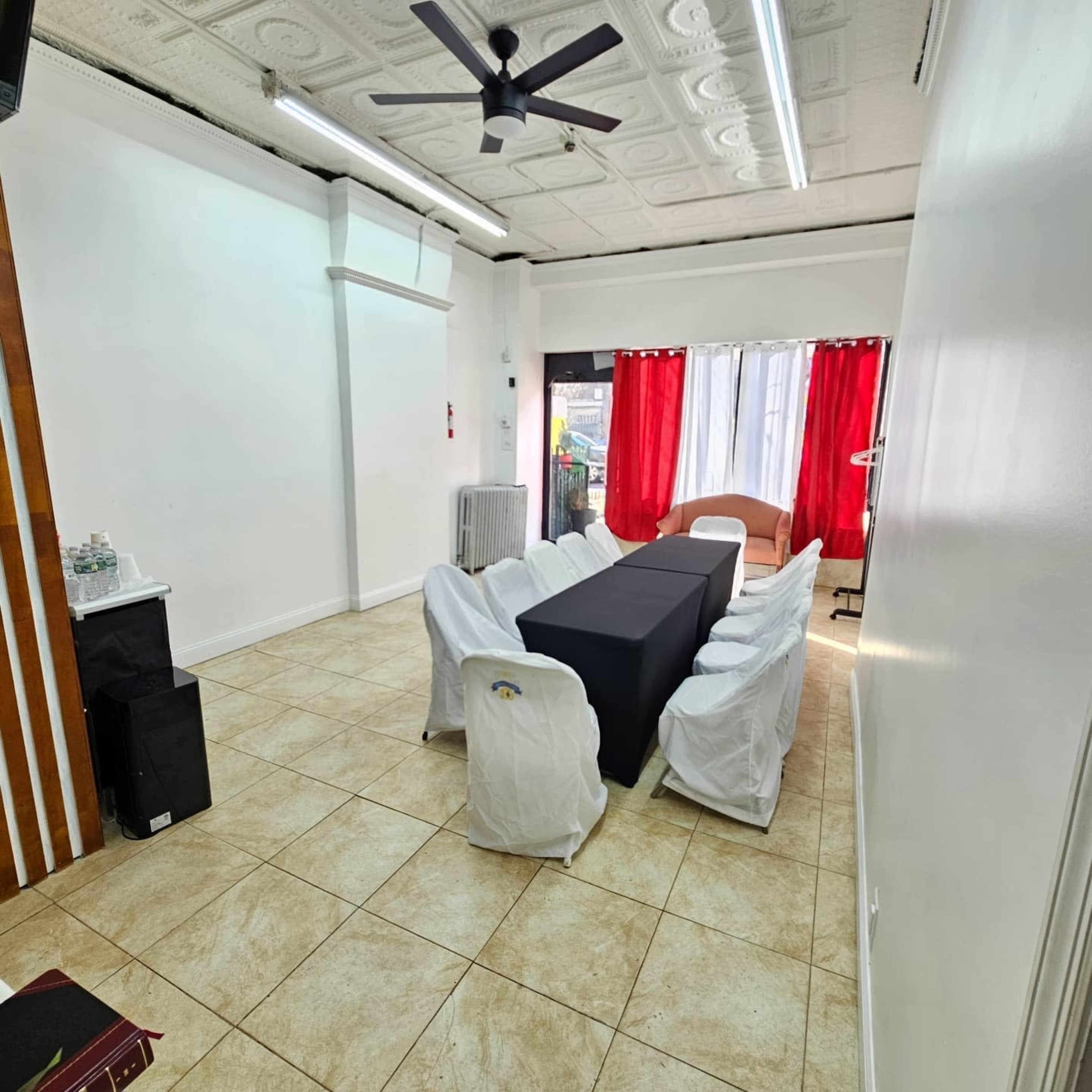 The room features a long table covered with a dark tablecloth, surrounded by white chairs, and is illuminated by natural light through a window with red curtains.
