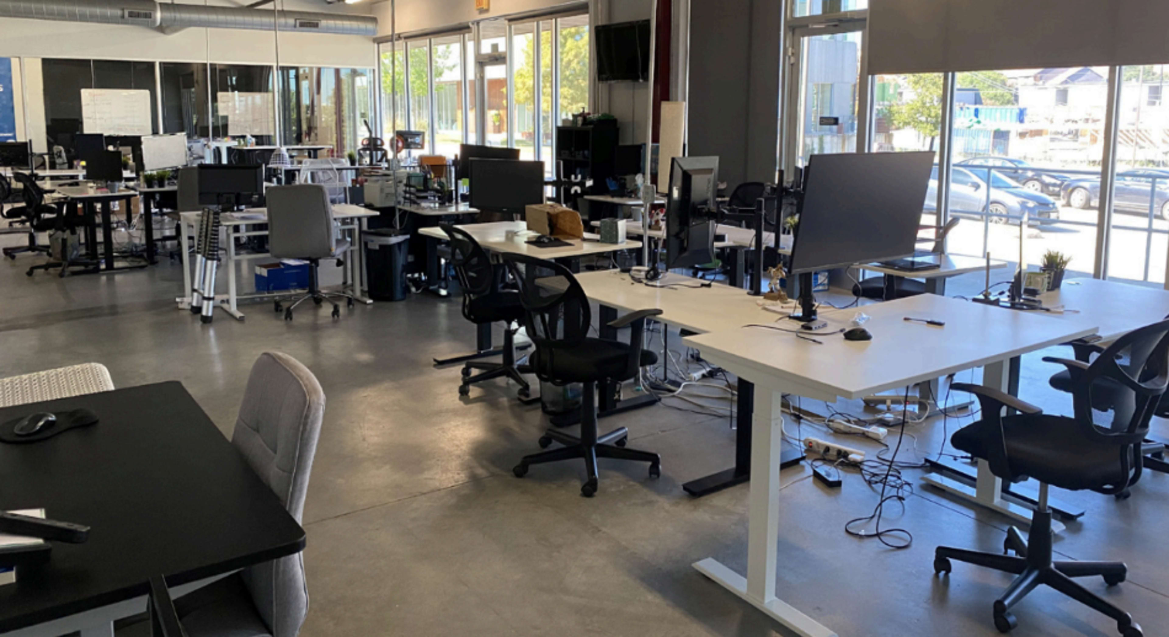 The image shows a modern open office space with multiple desks, computer monitors, and office chairs arranged in a bright, spacious environment.
