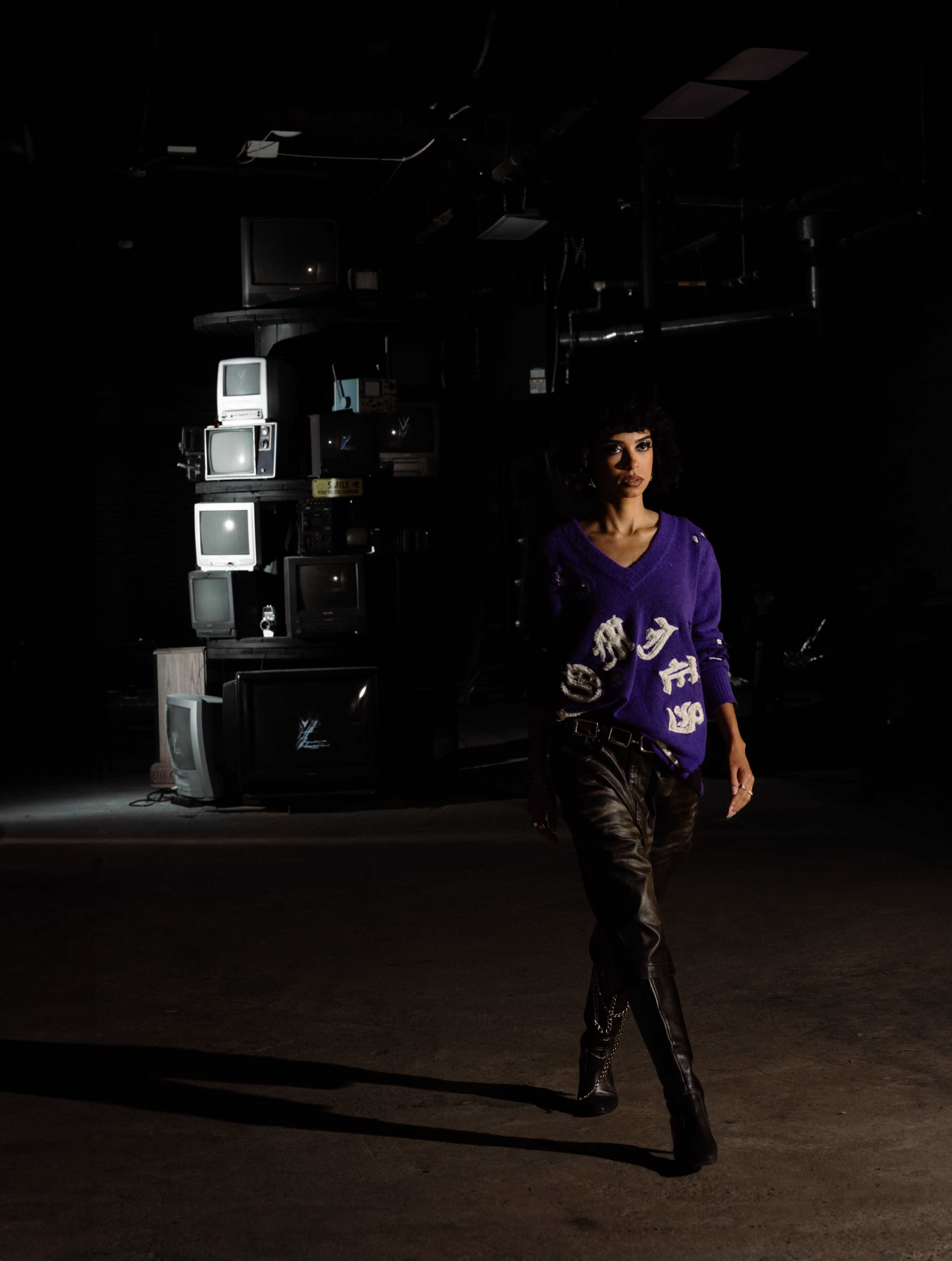 A model in a purple sweater and black leather pants walks past a stack of vintage televisions in a dimly lit industrial space.