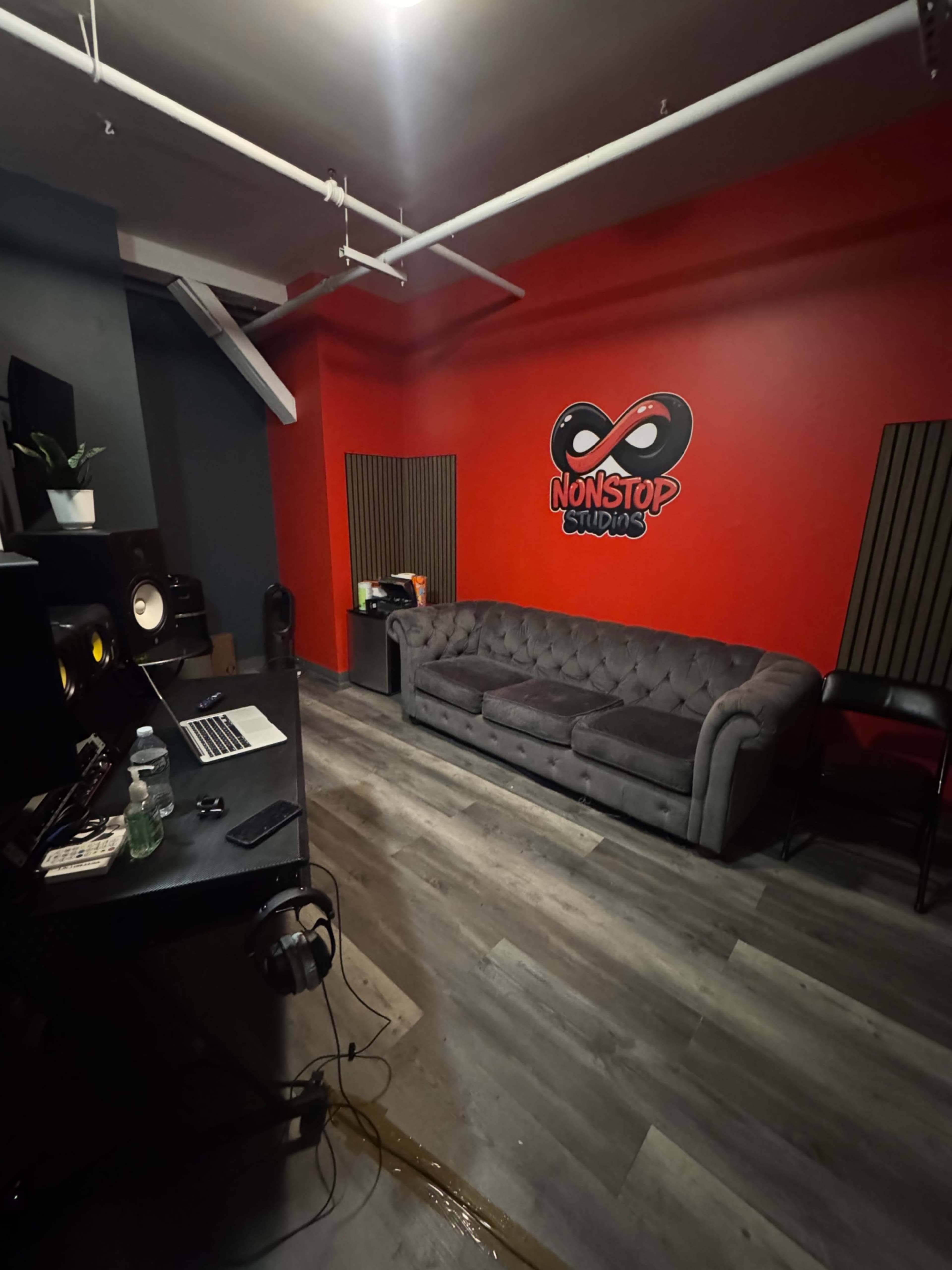 The image shows a recording studio with a red accent wall featuring the "NONSTOP STUDIOS" logo, a gray couch, and soundproof panels.