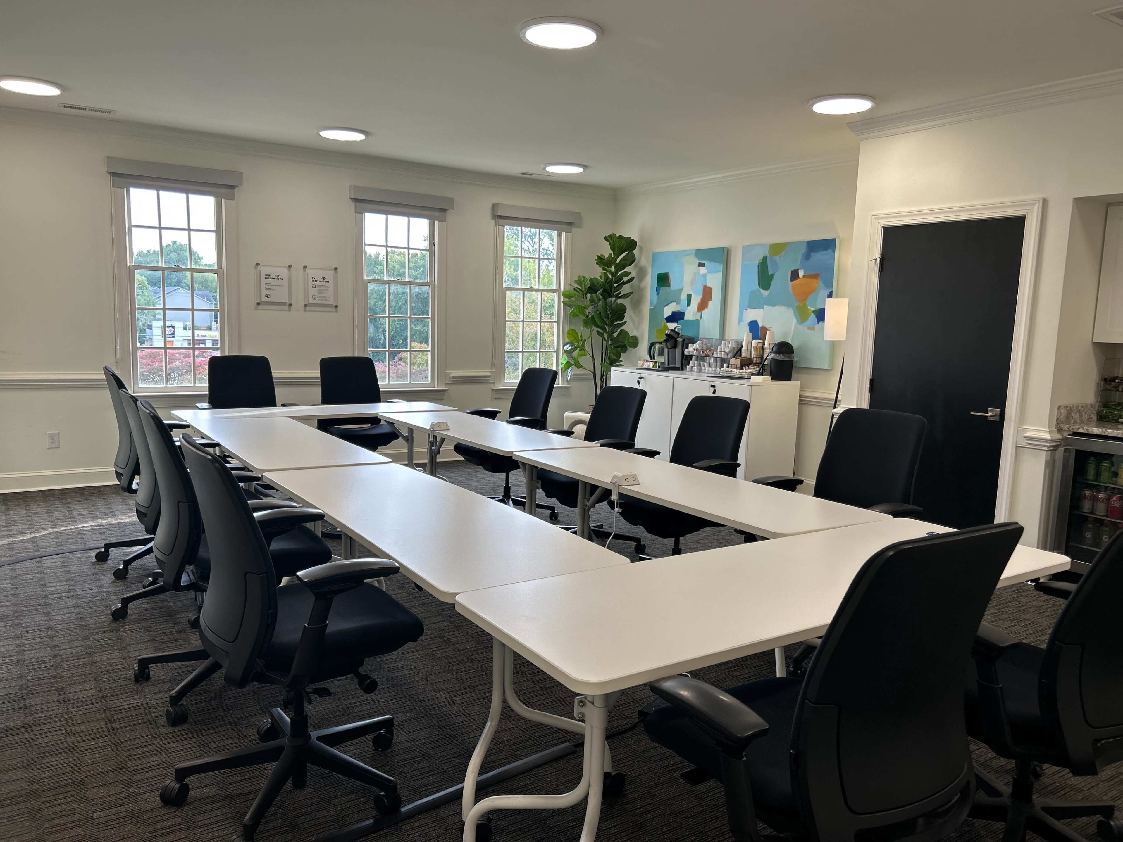 A modern conference room features a long white table surrounded by black ergonomic chairs, with large windows providing natural light and a refreshment area in the background.