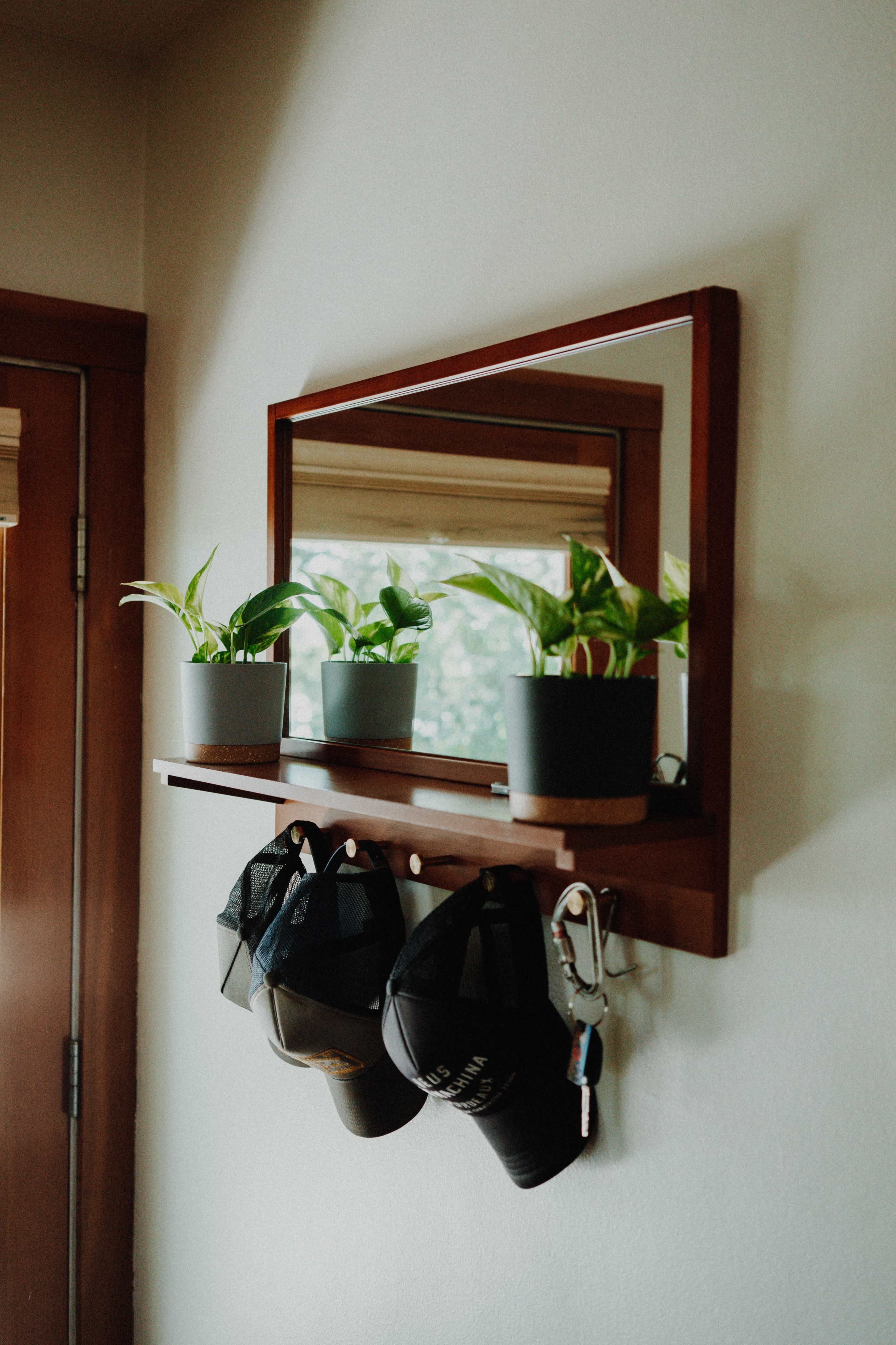 A mirror mounted above a shelf displays three potted plants and several hats hanging below.