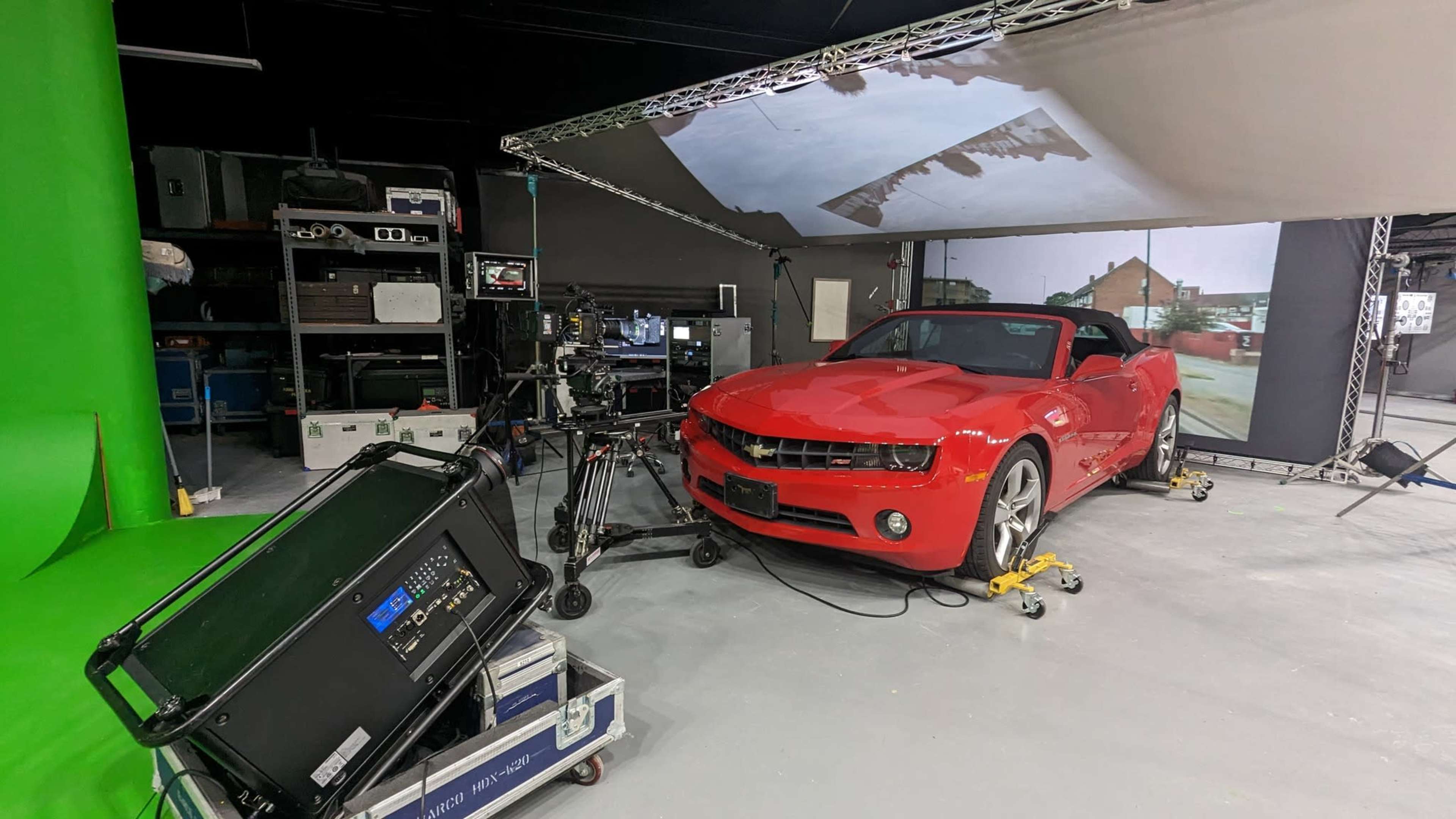 A red convertible car is positioned in a studio with a green screen and large backdrop displaying an outdoor scene.