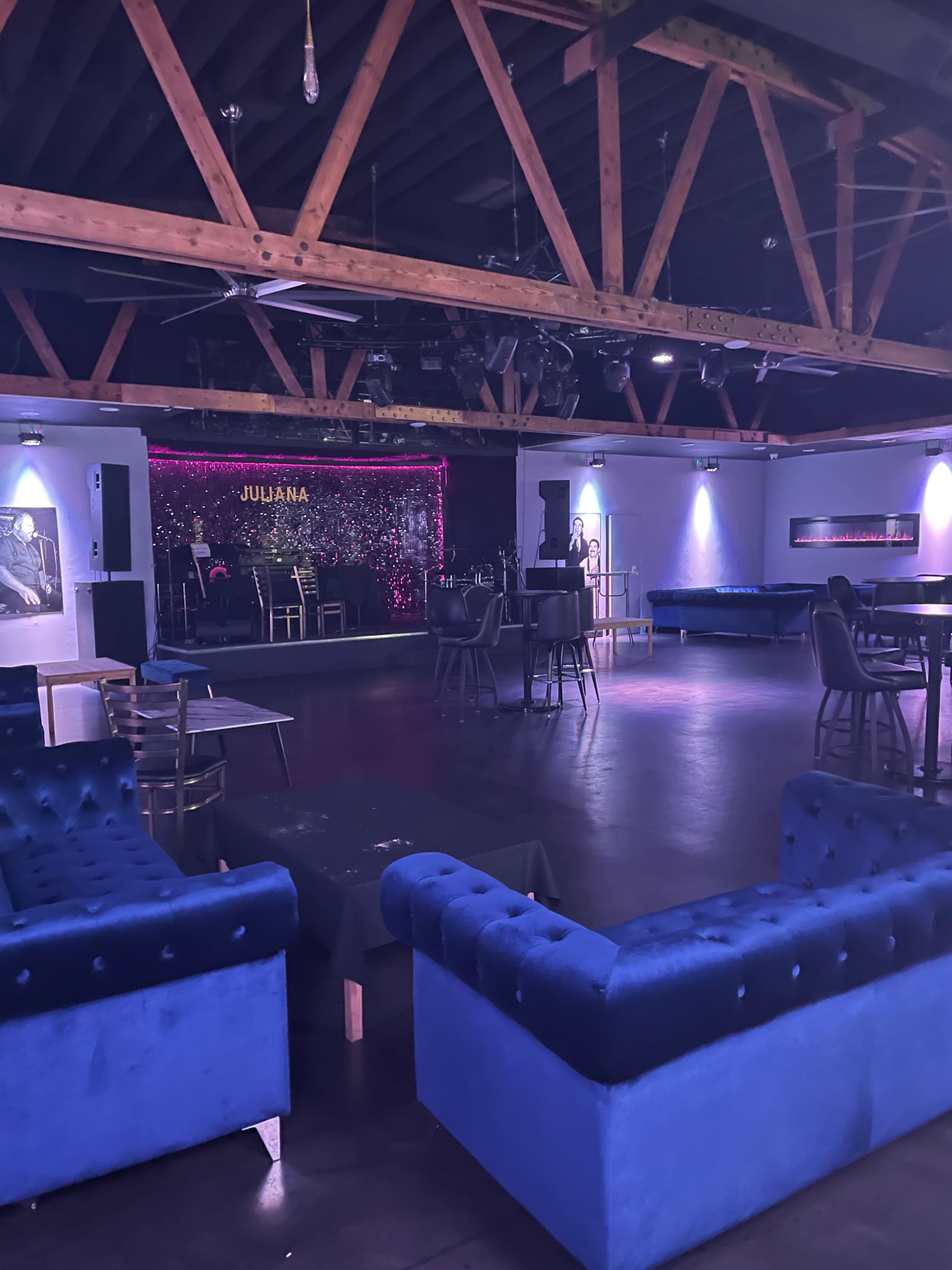 An empty lounge with blue upholstered seating, wooden beams, and a stage lit with purple lights in the background.