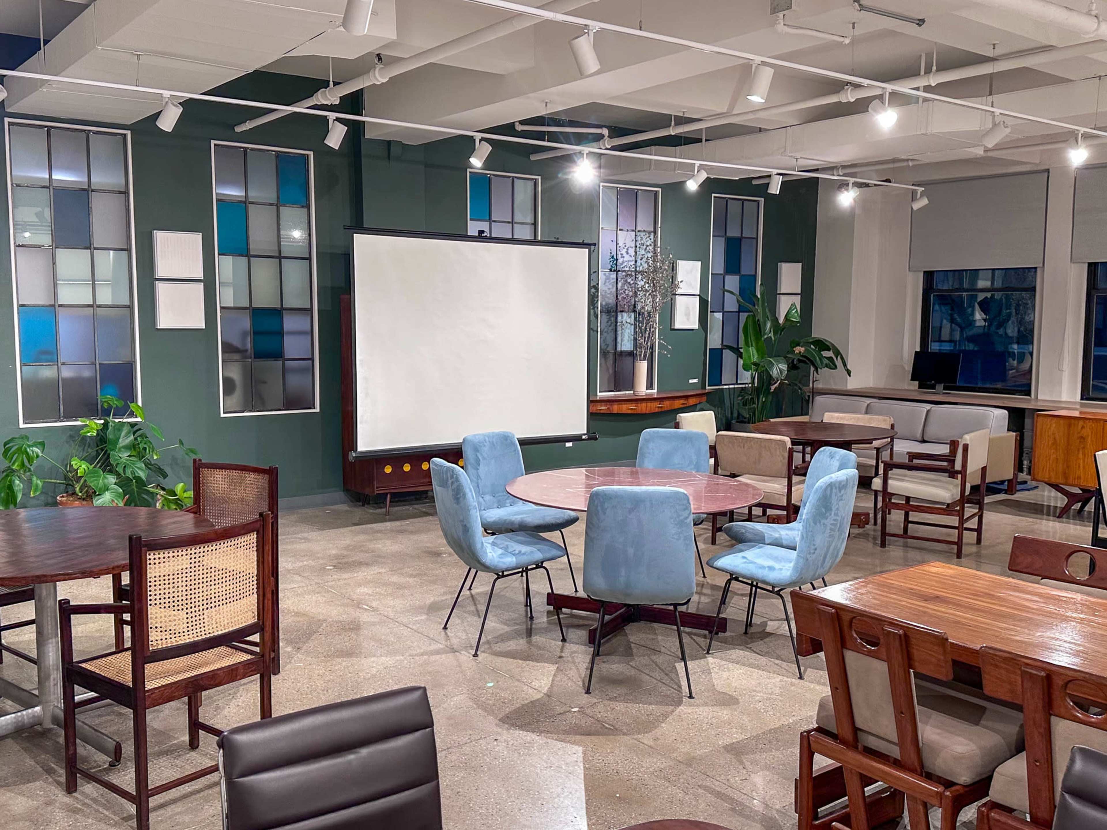 The image shows a modern workspace with several round and rectangular tables surrounded by chairs, a large projection screen, and plants in a bright room.