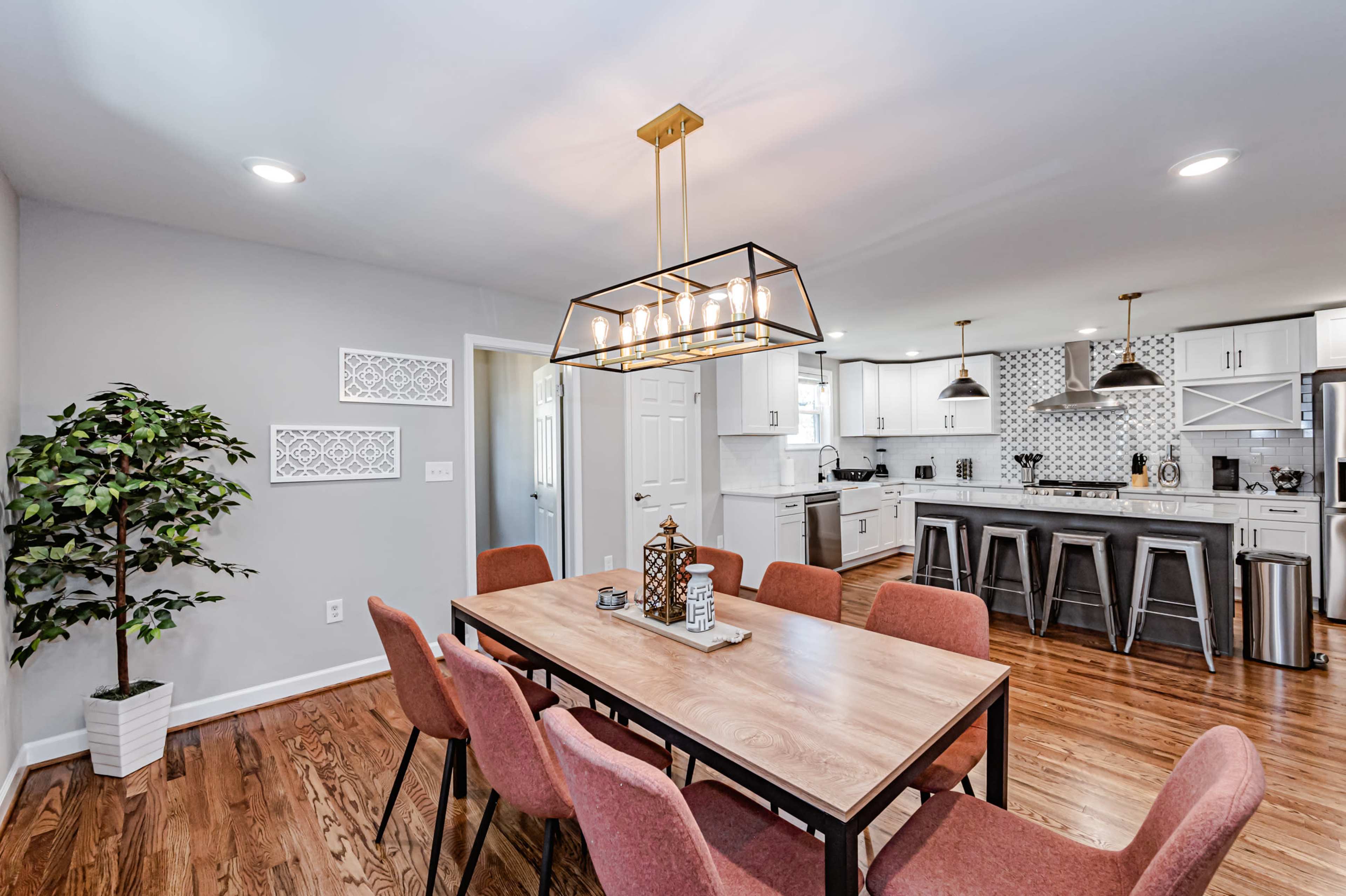 A modern dining area features a wooden table surrounded by pink chairs, with a large light fixture and a view into a bright kitchen.