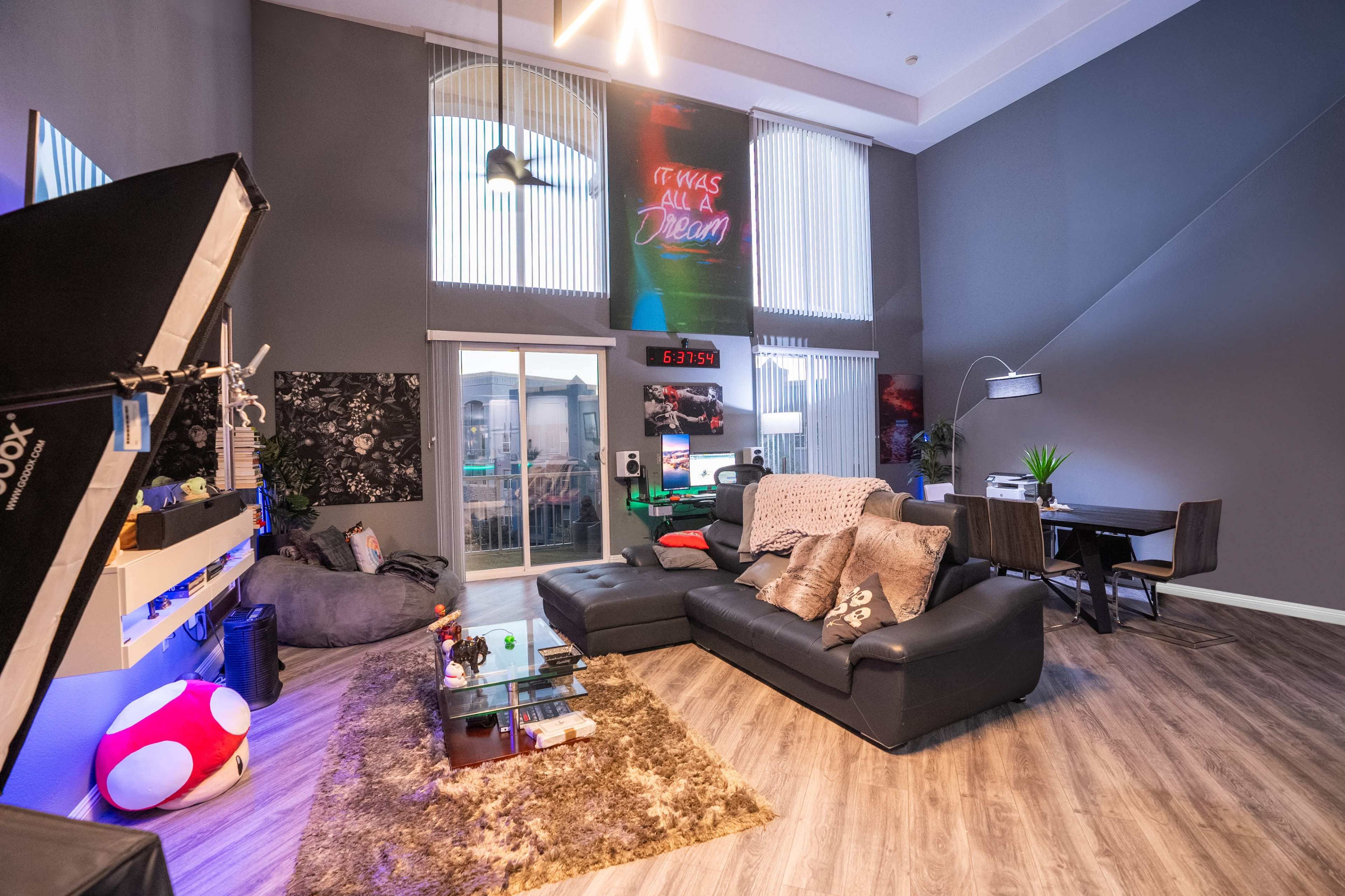 A contemporary living room featuring black leather sofas, a large area rug, and a neon sign on the wall, with large windows providing ample natural light.