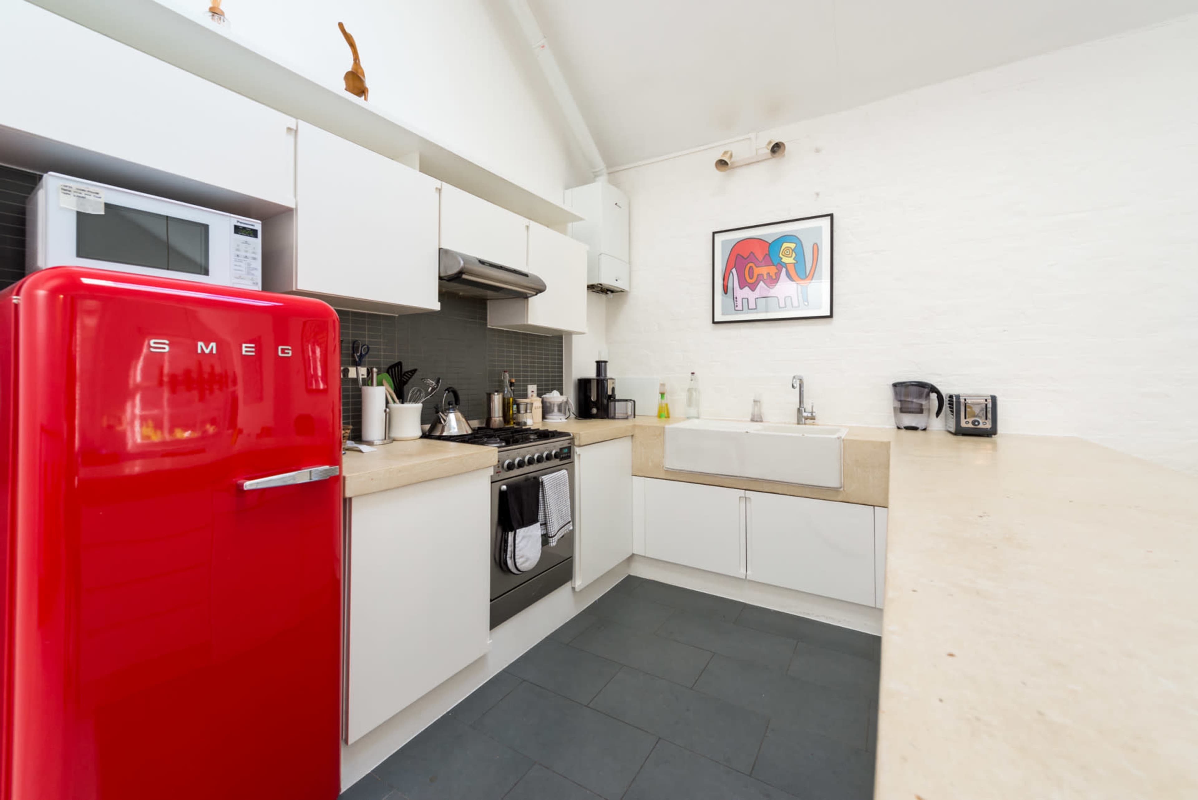 The image shows a modern kitchen featuring a red Smeg refrigerator, white cabinetry, a stainless steel oven, and a farmhouse sink.