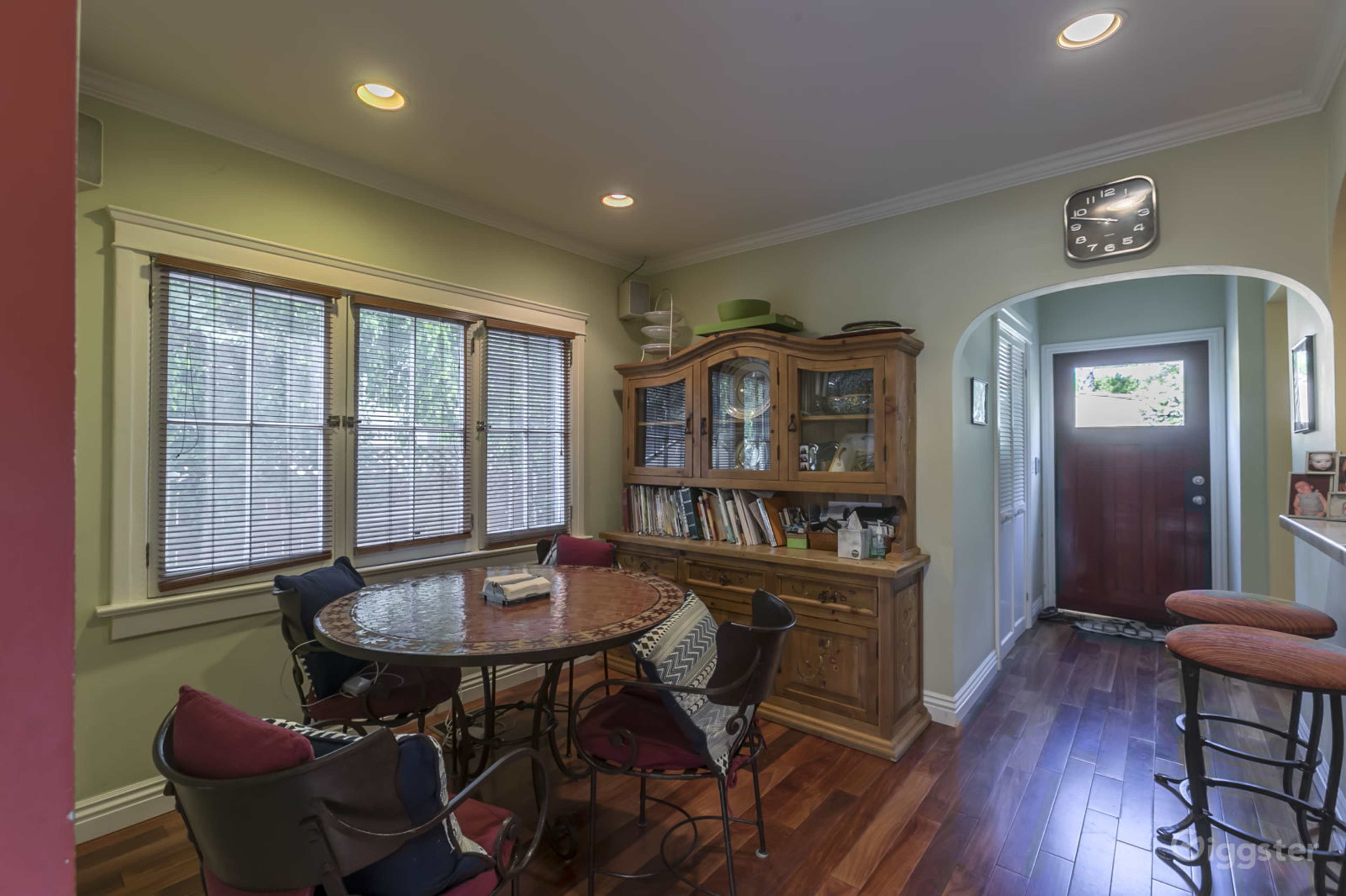 The image shows a dining area with a round table surrounded by chairs, a wooden cabinet displaying books, and a doorway leading to an entrance with a clock on the wall.