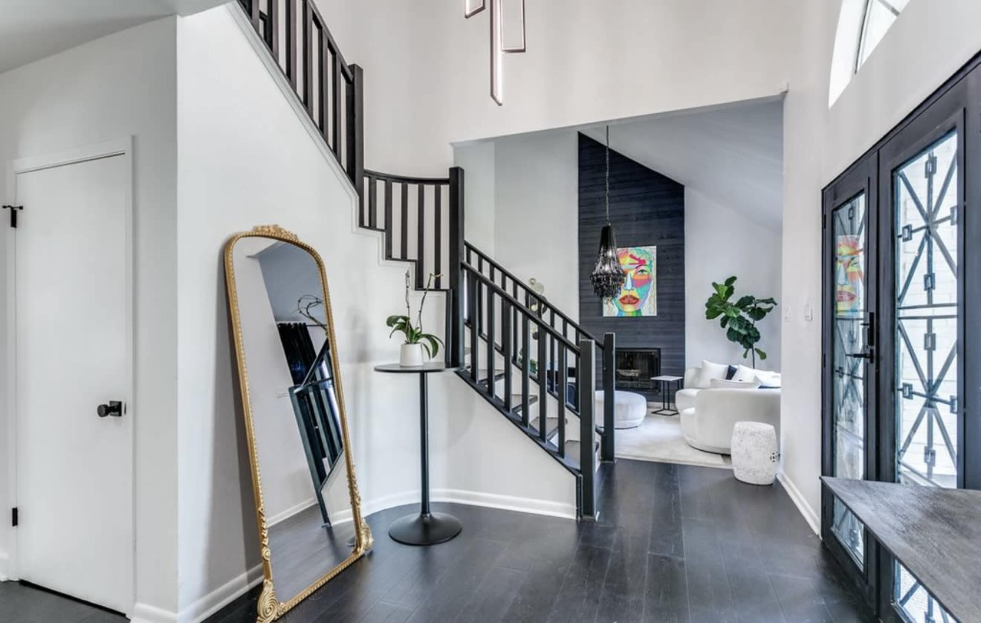 The image shows a modern entryway featuring a staircase, a large decorative mirror, and a cozy seating area visible in the background.