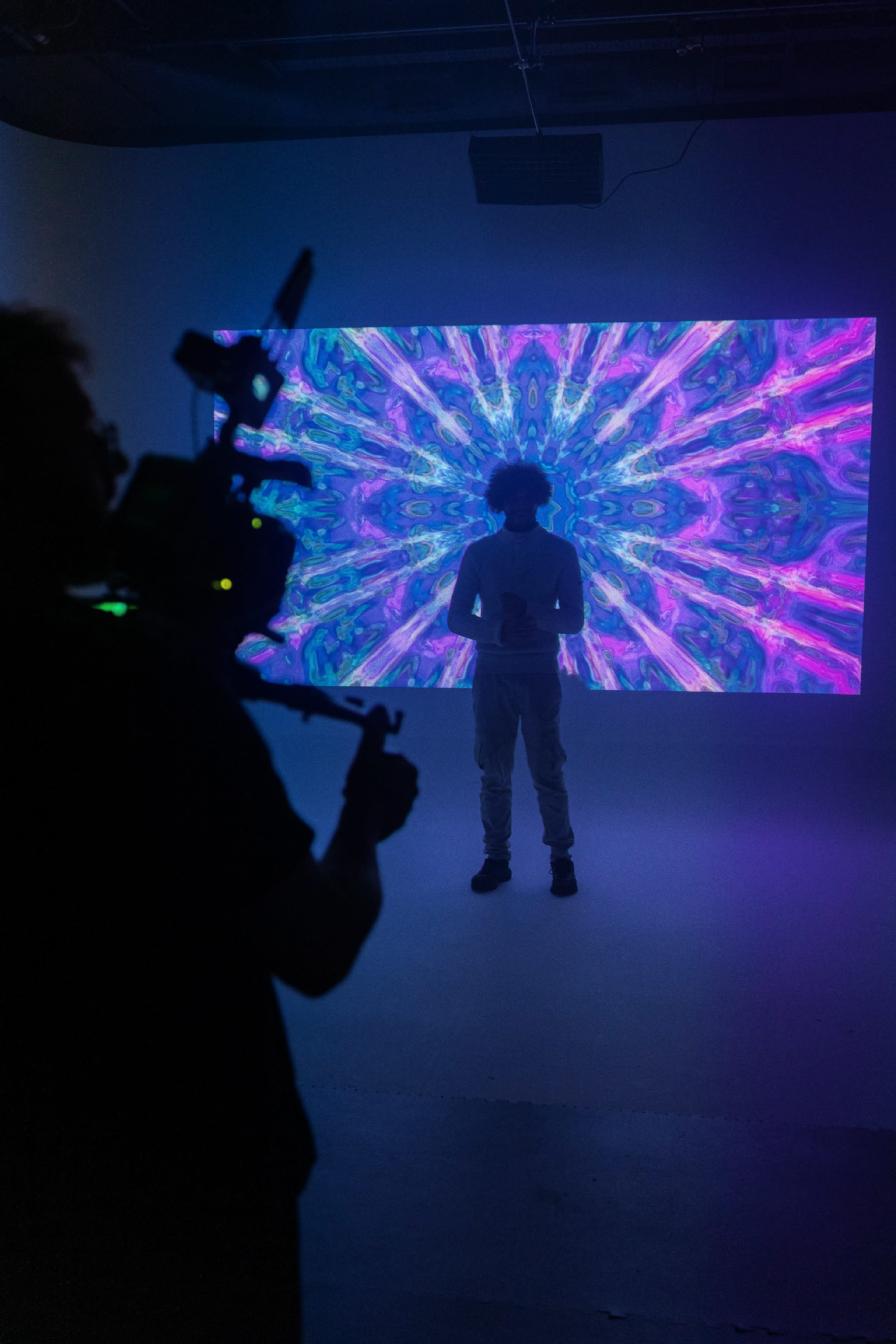 A person stands in front of a colorful, abstract projection on a wall while another individual films them with a camera.