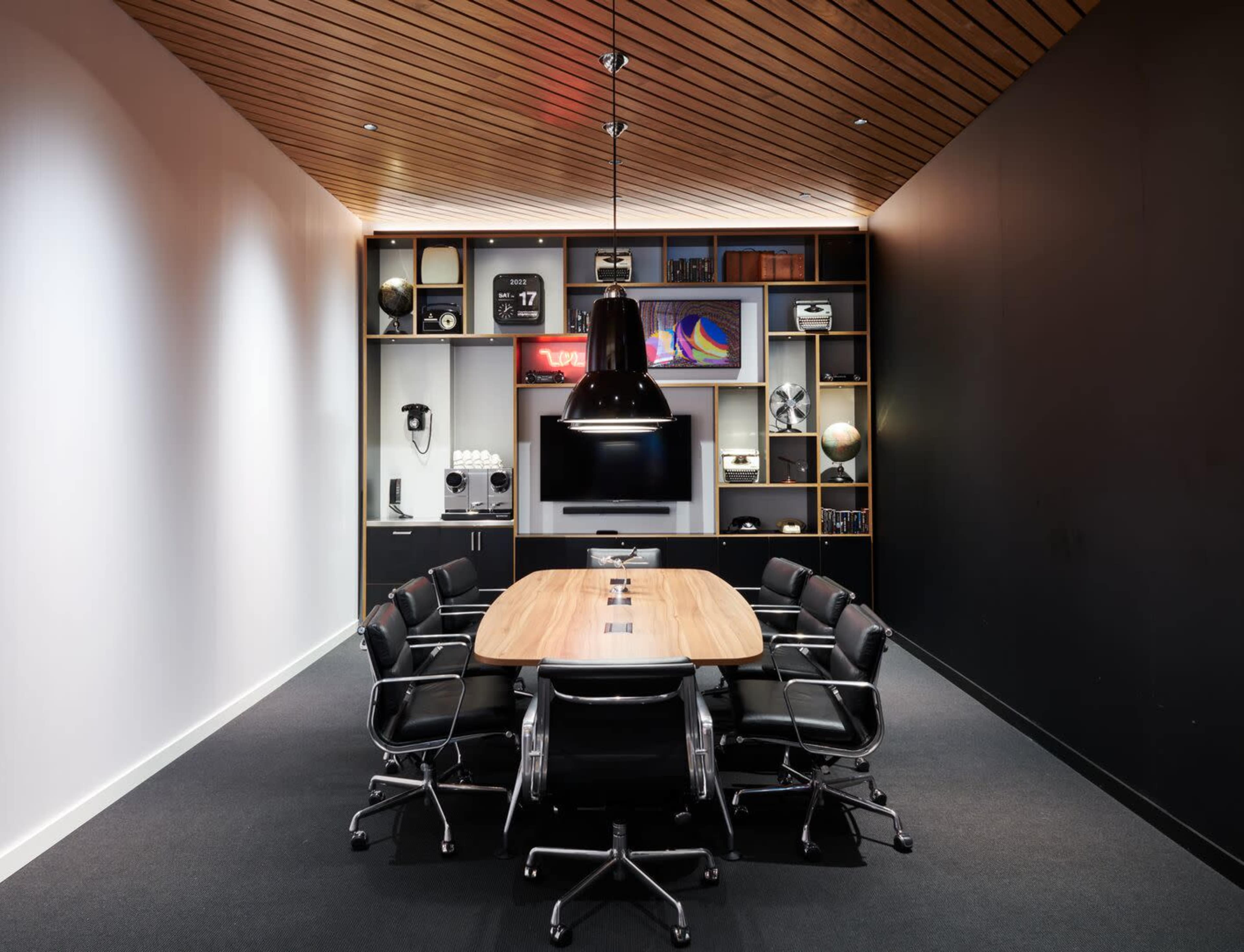 A modern conference room features a long wooden table surrounded by black chairs, with a multimedia setup and a wall of shelves displaying various items.