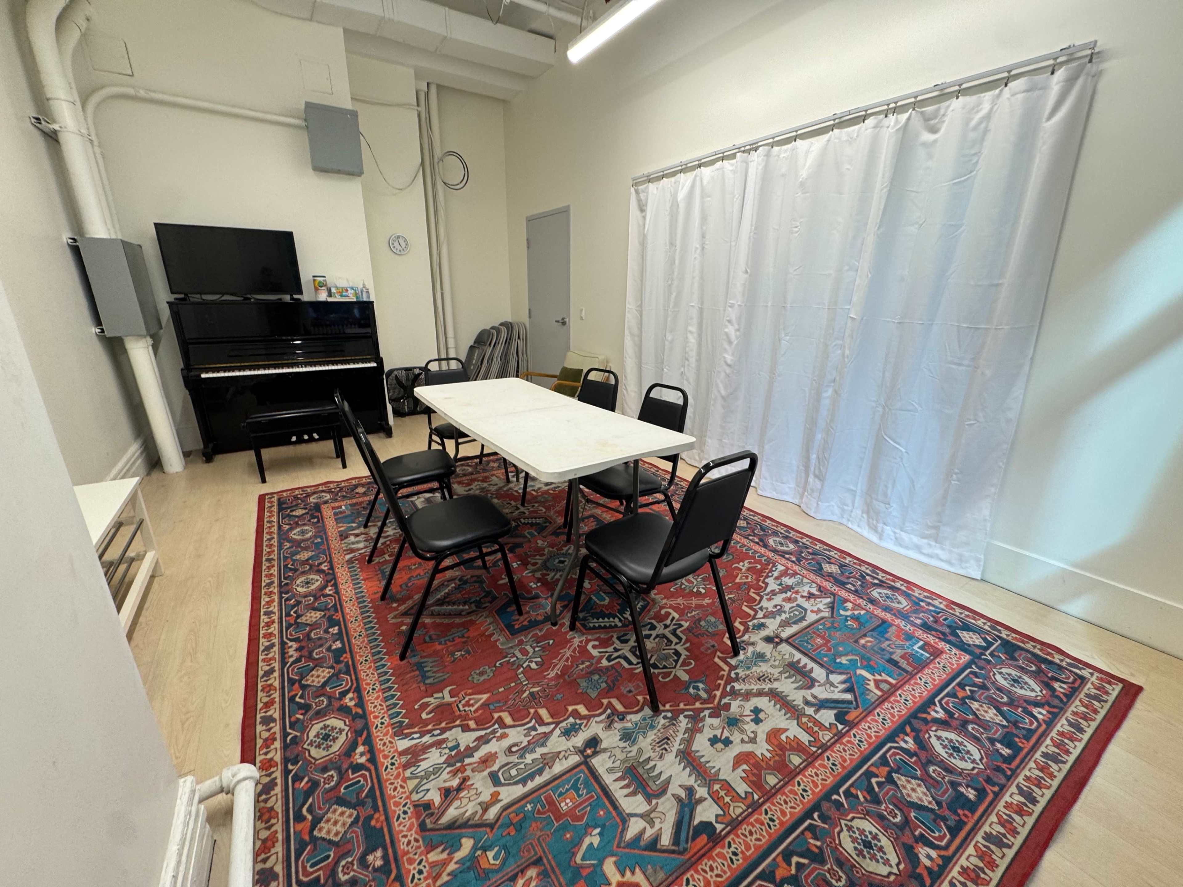The image shows a room with a table surrounded by six chairs, a piano in one corner, and a white curtain covering a section of the wall.