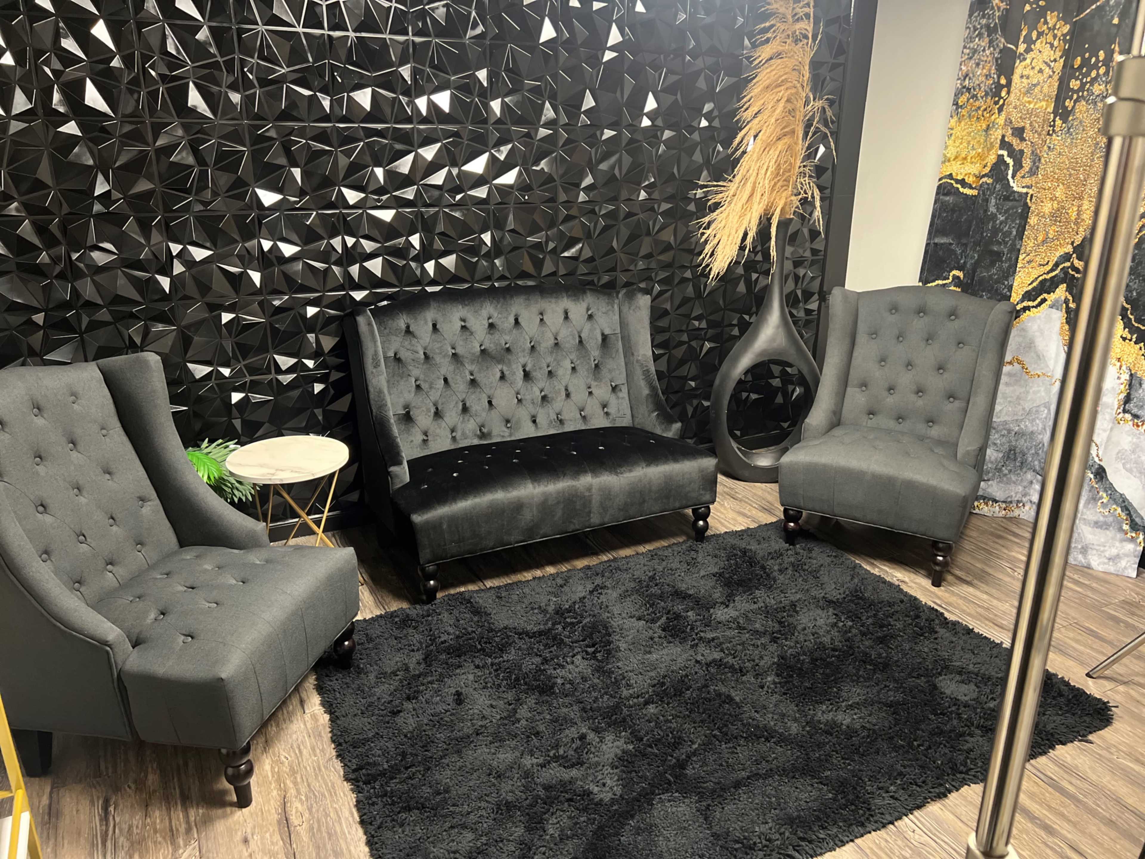 The image shows a stylish lounge area featuring tufted black chairs and a matching sofa arranged on a dark textured rug against a geometric black and gold wall.