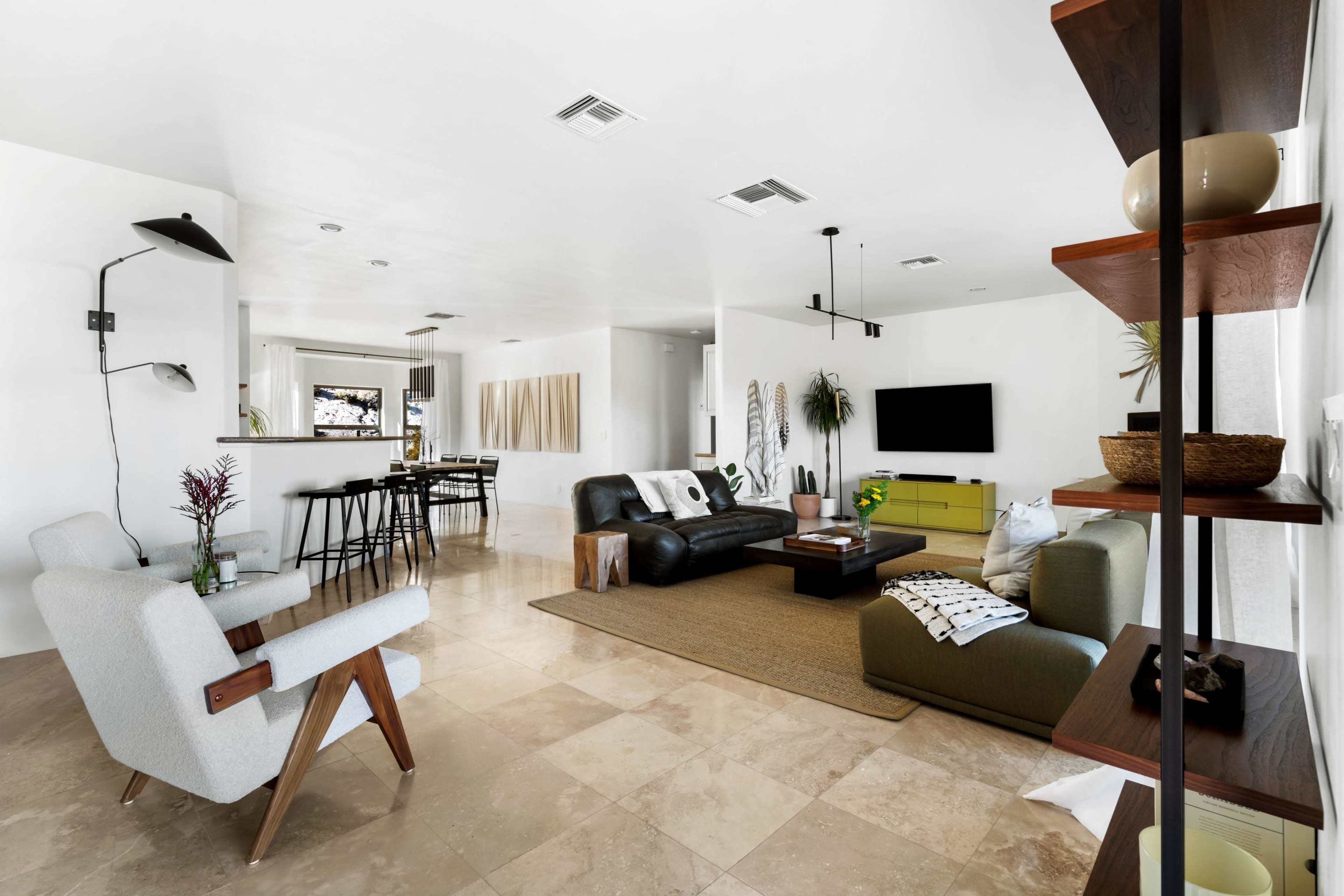 The image shows a modern living room featuring a black leather sofa, a green accent chair, and beige tile flooring, with a dining area visible in the background.