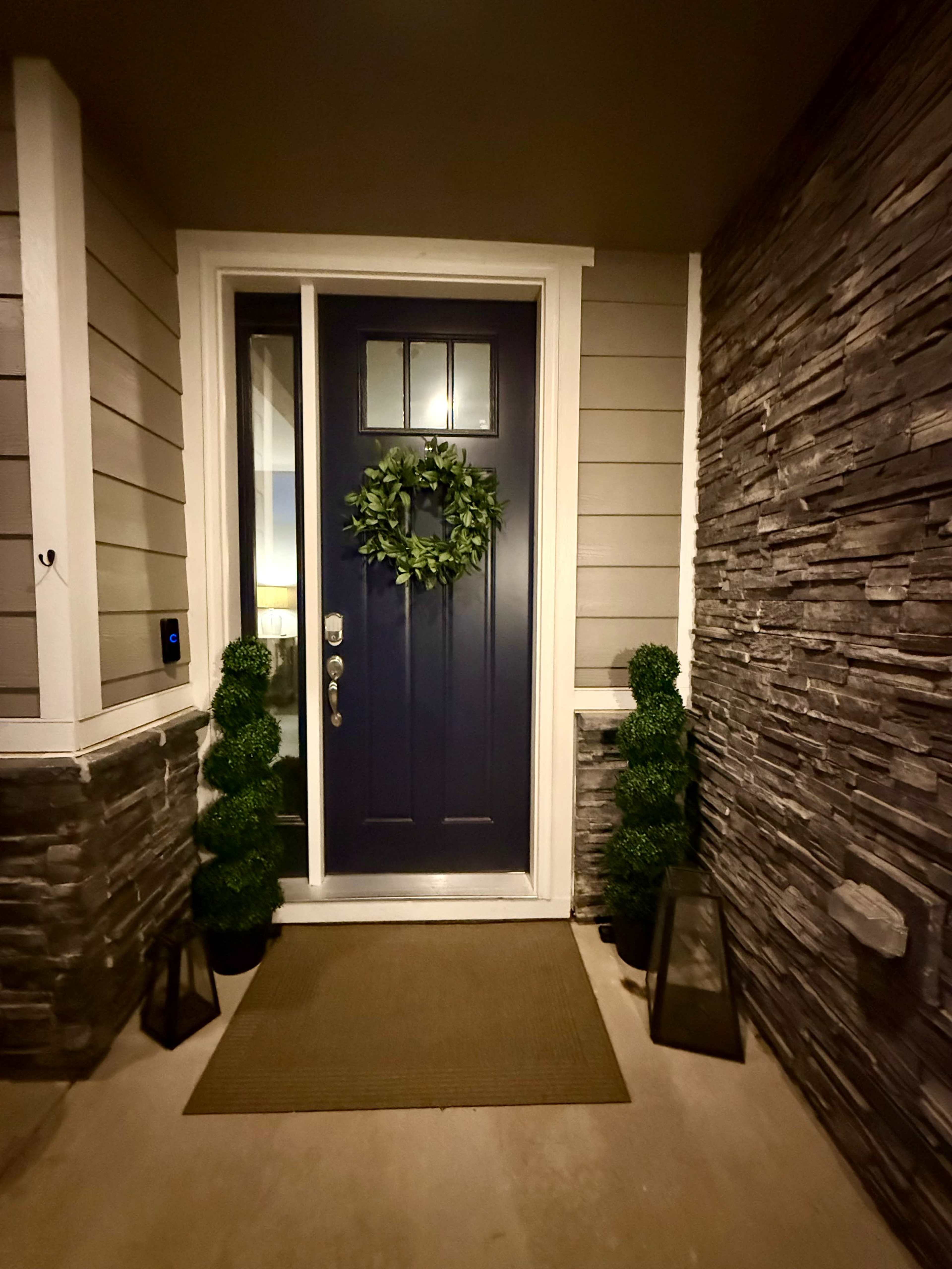 The image shows a front entryway with a dark blue door, a green wreath, two shaped green shrubs on either side, and stone accents on the wall.