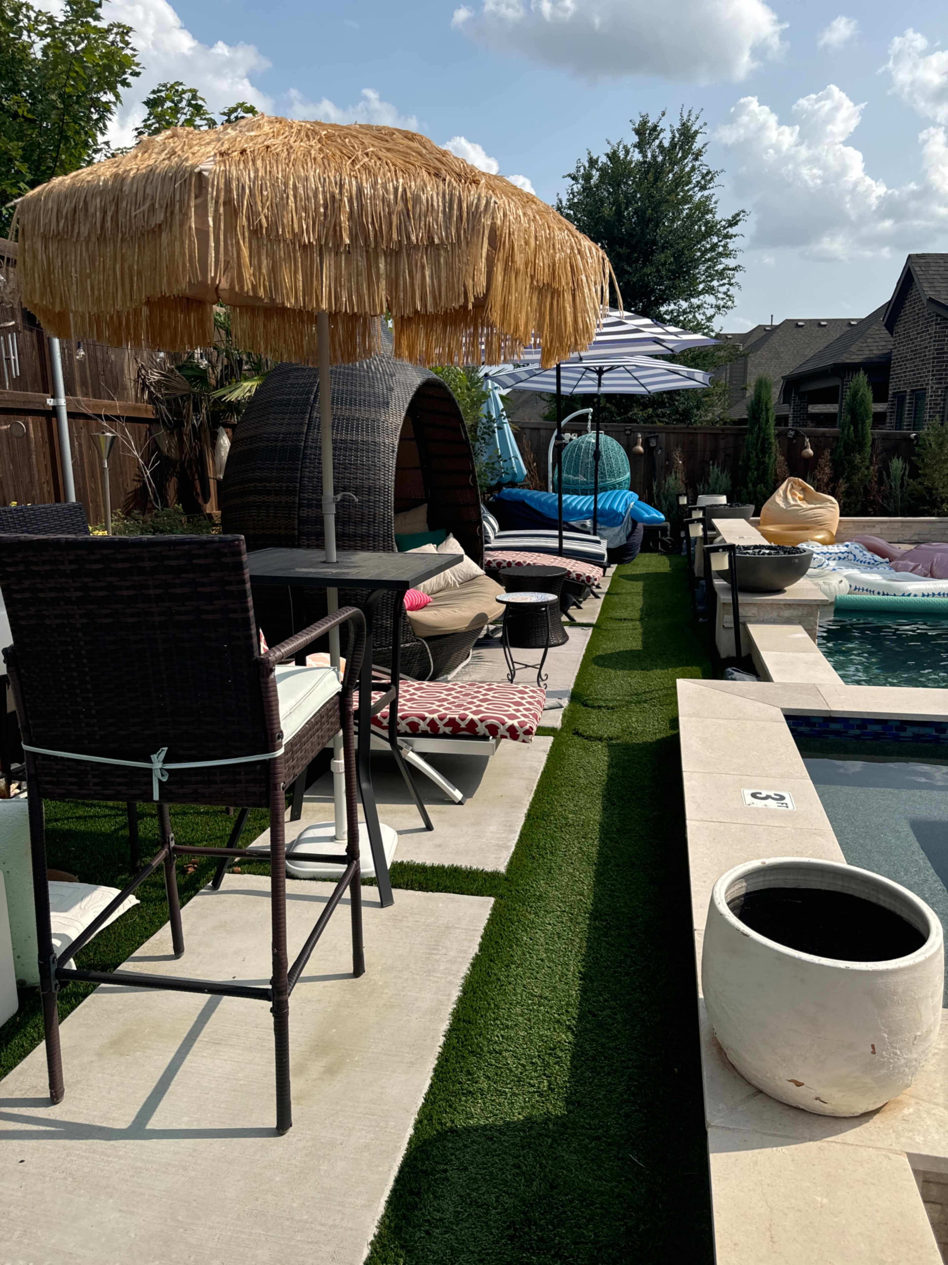 The scene shows a backyard patio area featuring a straw-topped umbrella, lounge seating, a small pool with surrounding tiles, and a green artificial grass walkway.