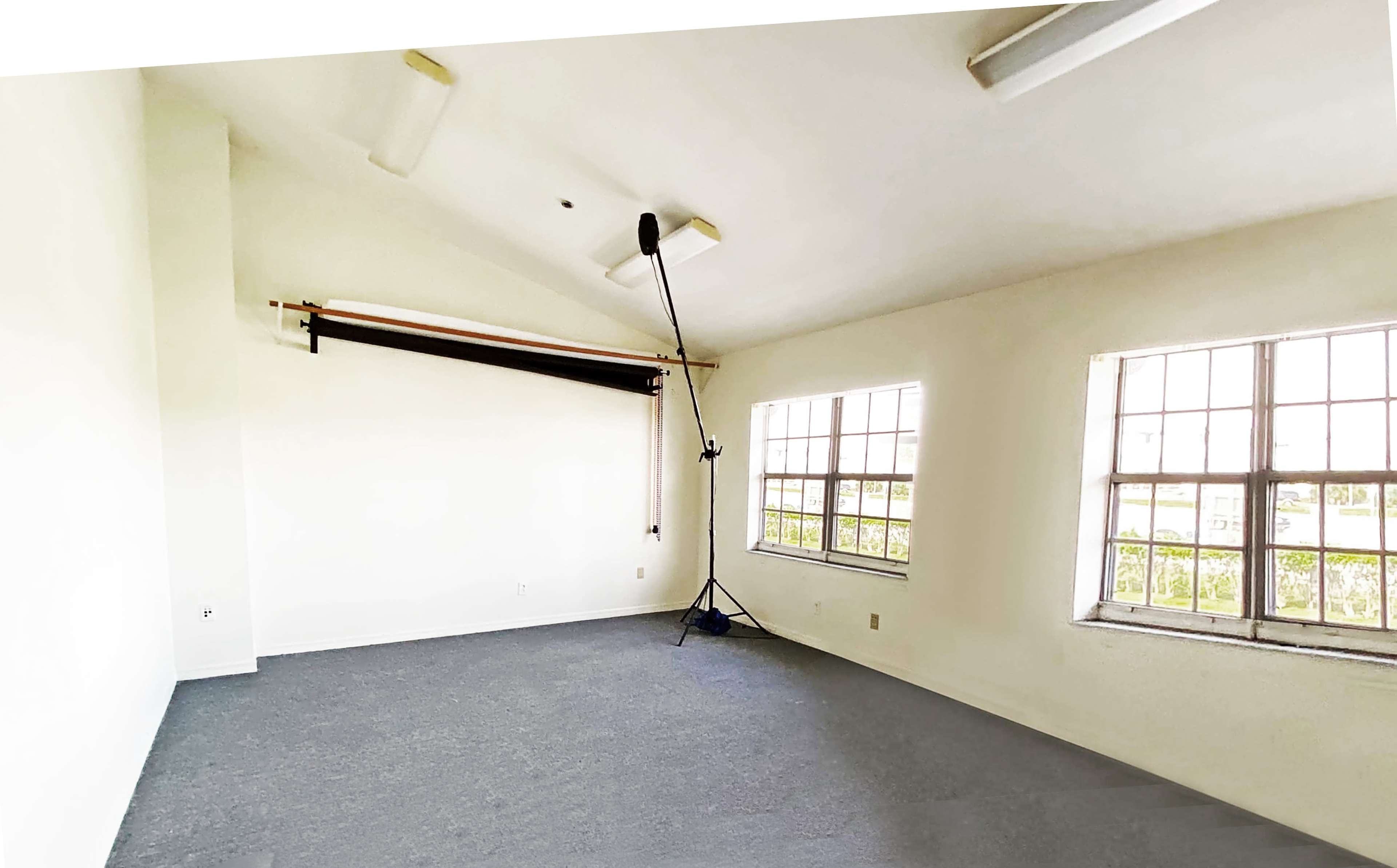 The image shows a spacious, empty room with two windows, a gray floor, and a backdrop support stand mounted on the ceiling.