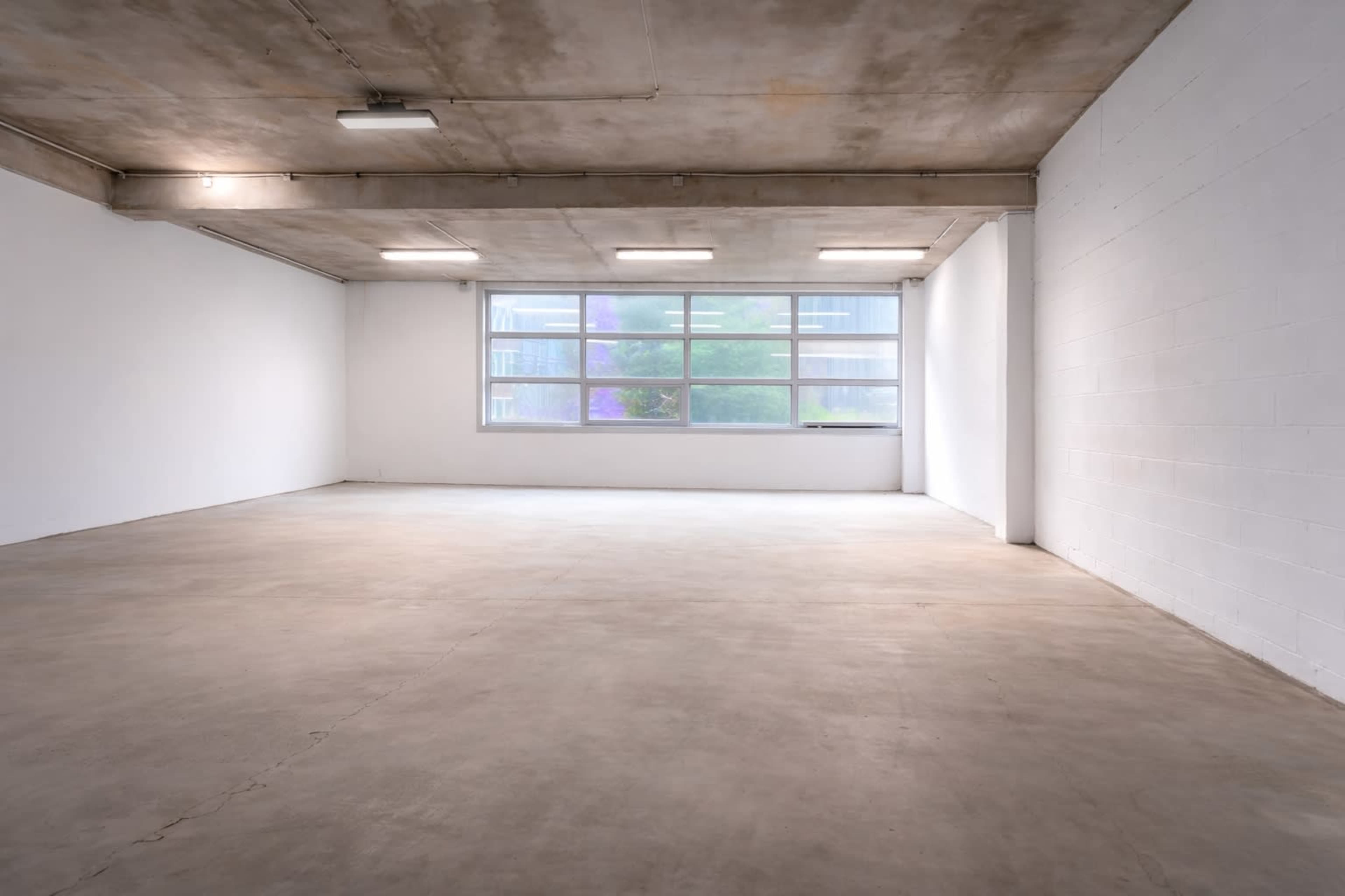 The image shows a large, empty room with a concrete floor and a window allowing natural light to enter.