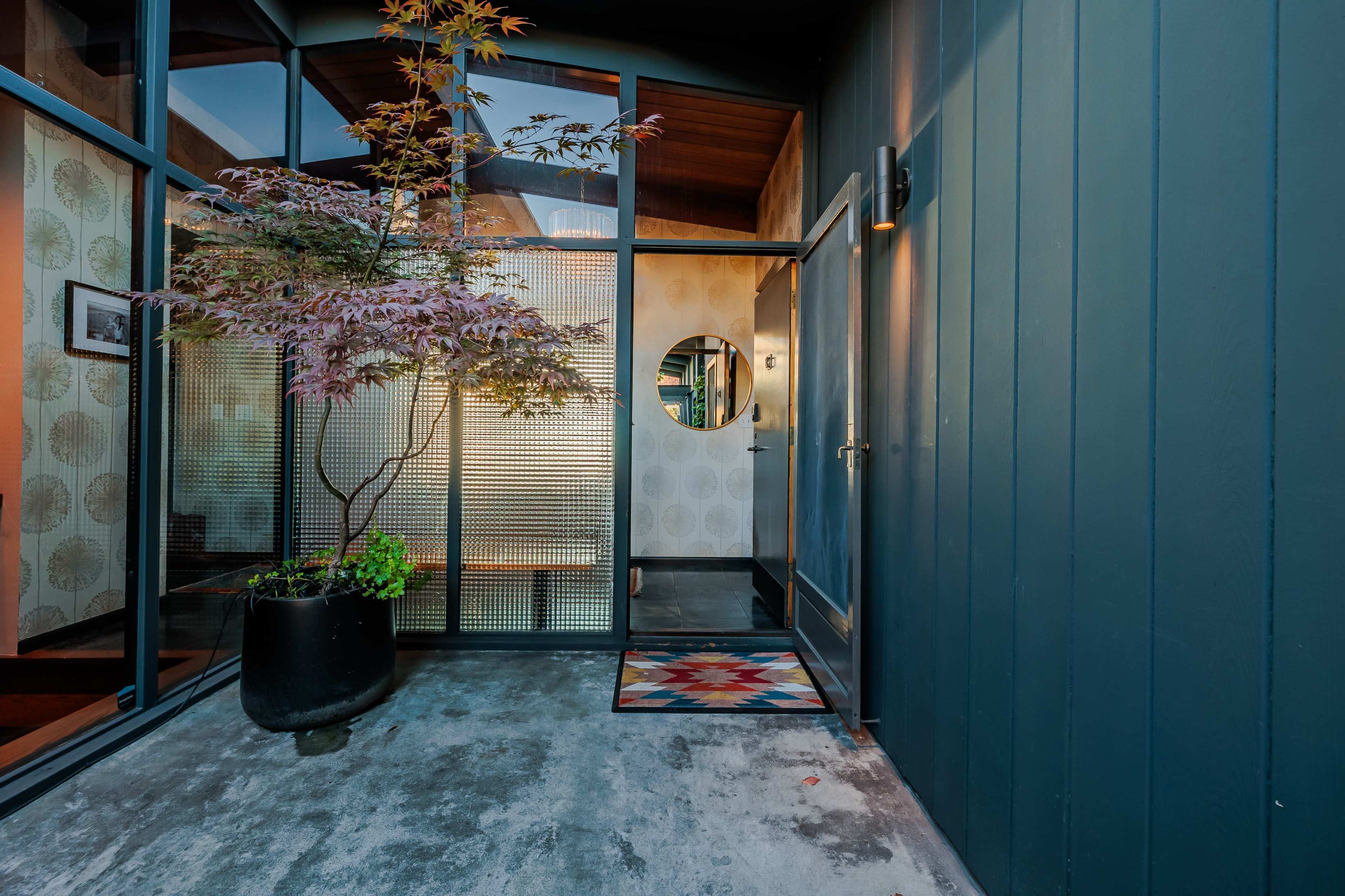 The entrance features a modern design with a circular window, a potted tree, and a patterned welcome mat on a concrete floor.