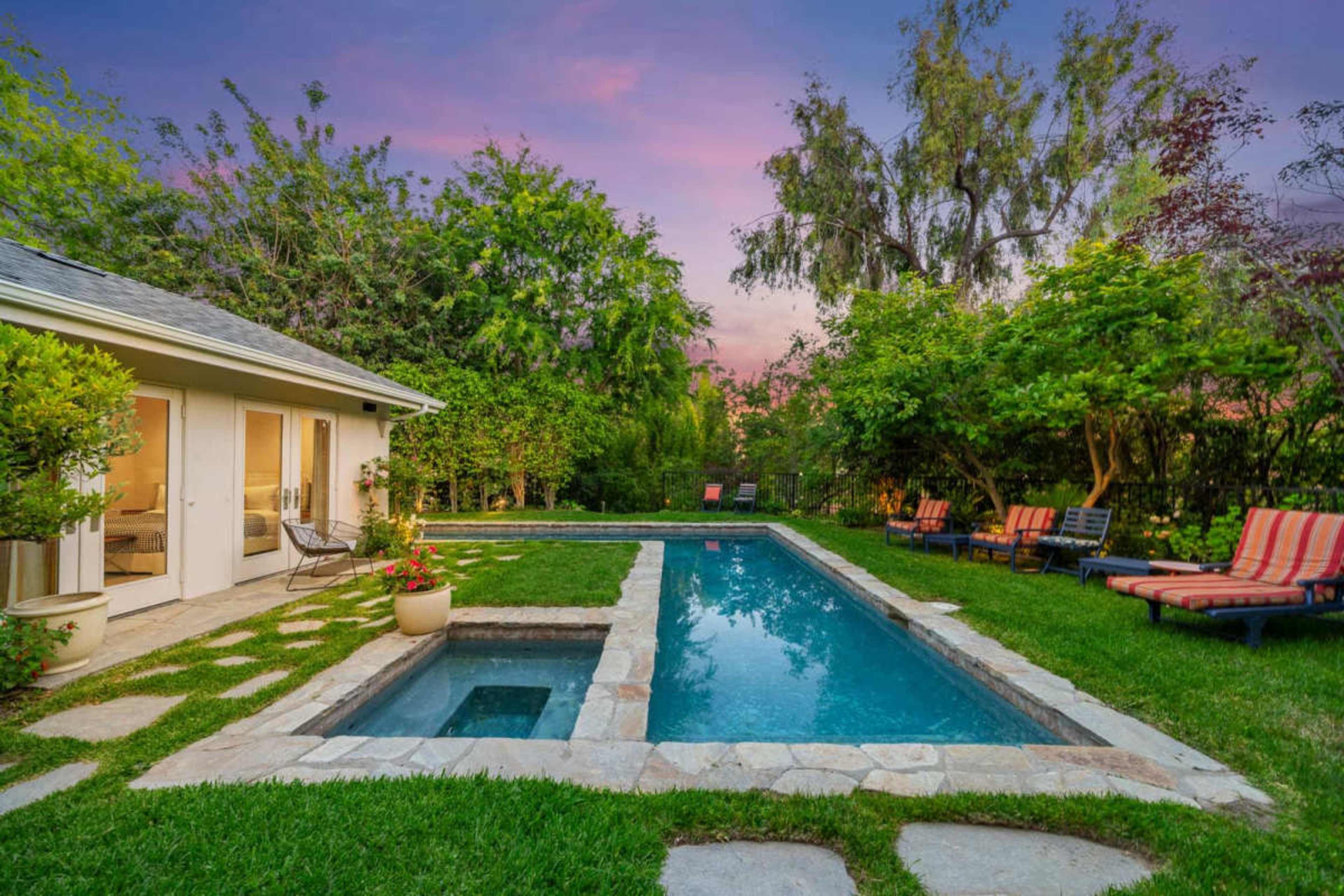 The image shows a backyard with a swimming pool, spa, and seating area surrounded by greenery and a house in the background.