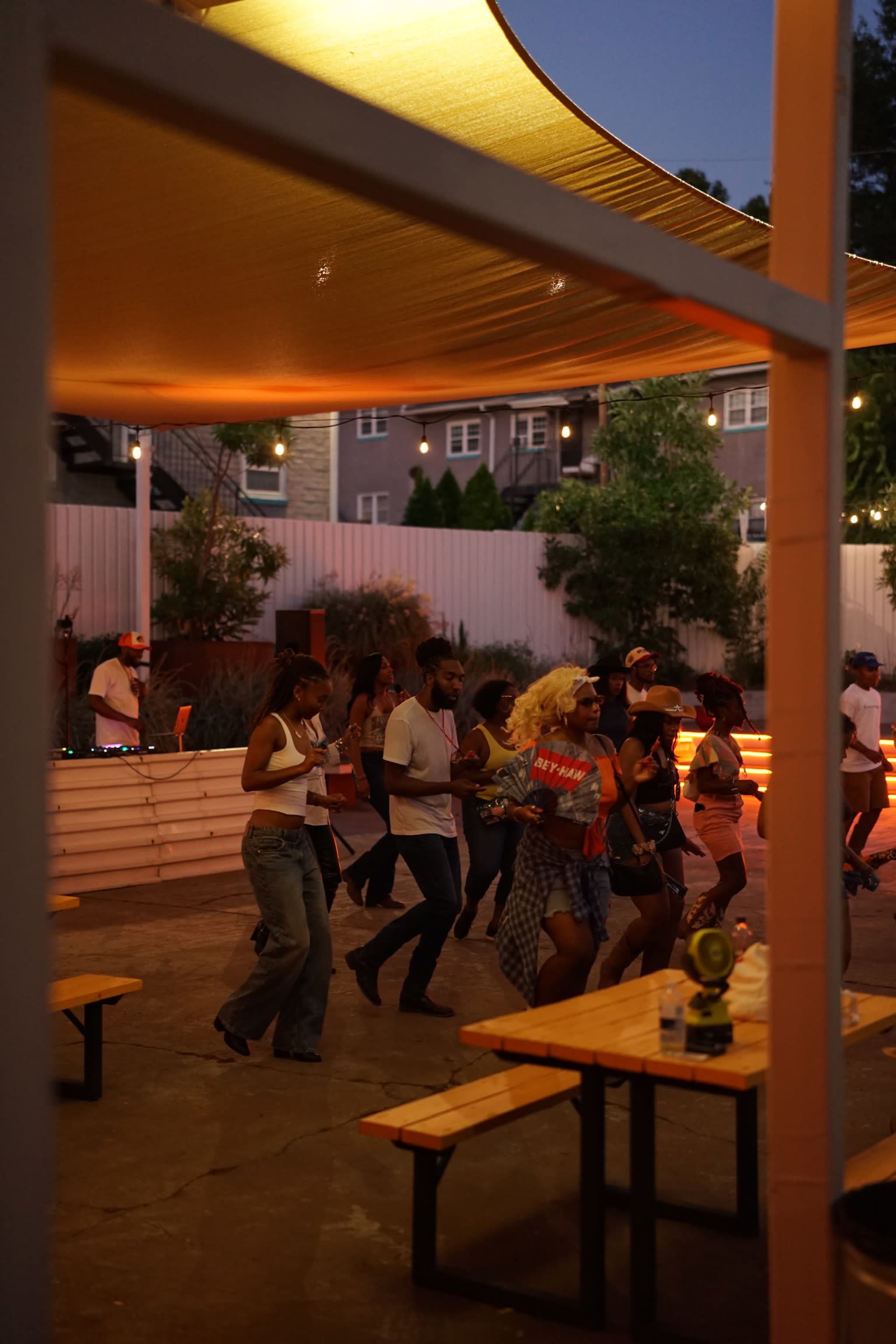 A group of people are dancing under a large canopy in an outdoor venue, with string lights illuminating the space.