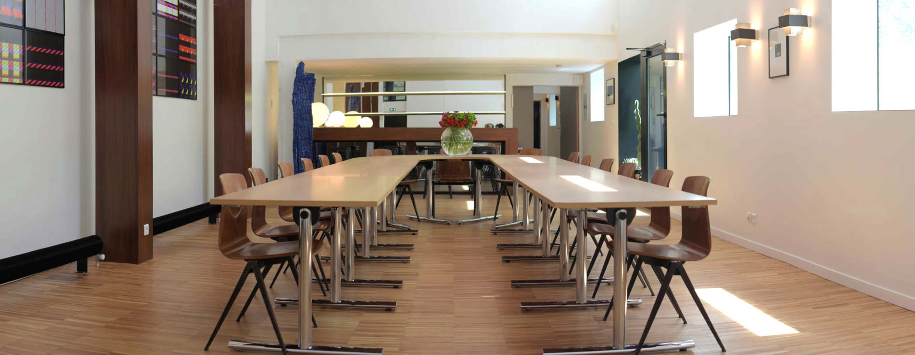 A spacious conference room features a long table set with chairs, illuminated by natural light from large windows.