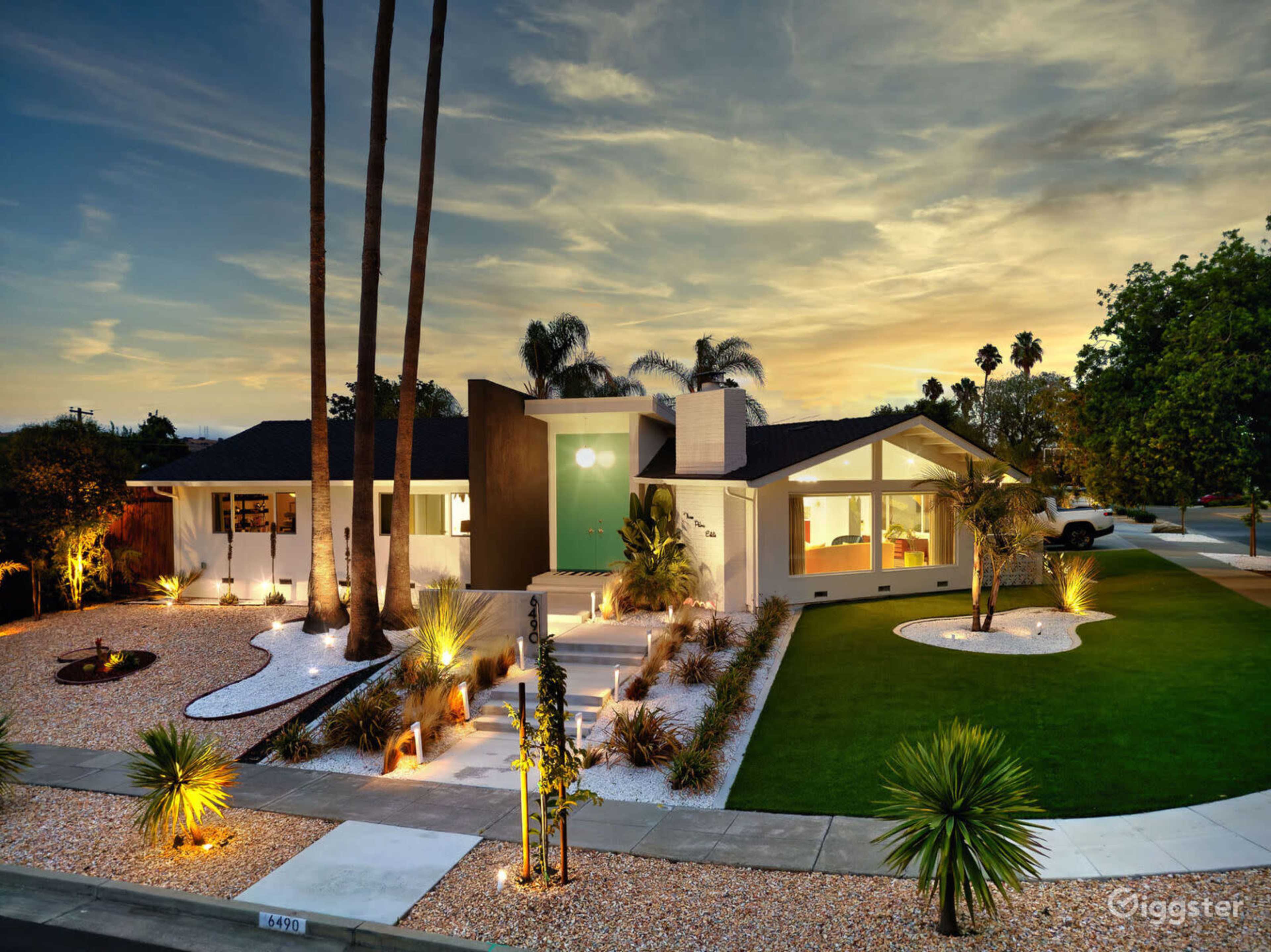 A modern single-story house features a landscaped front yard with palm trees, gravel pathways, and a well-lit entrance at dusk.