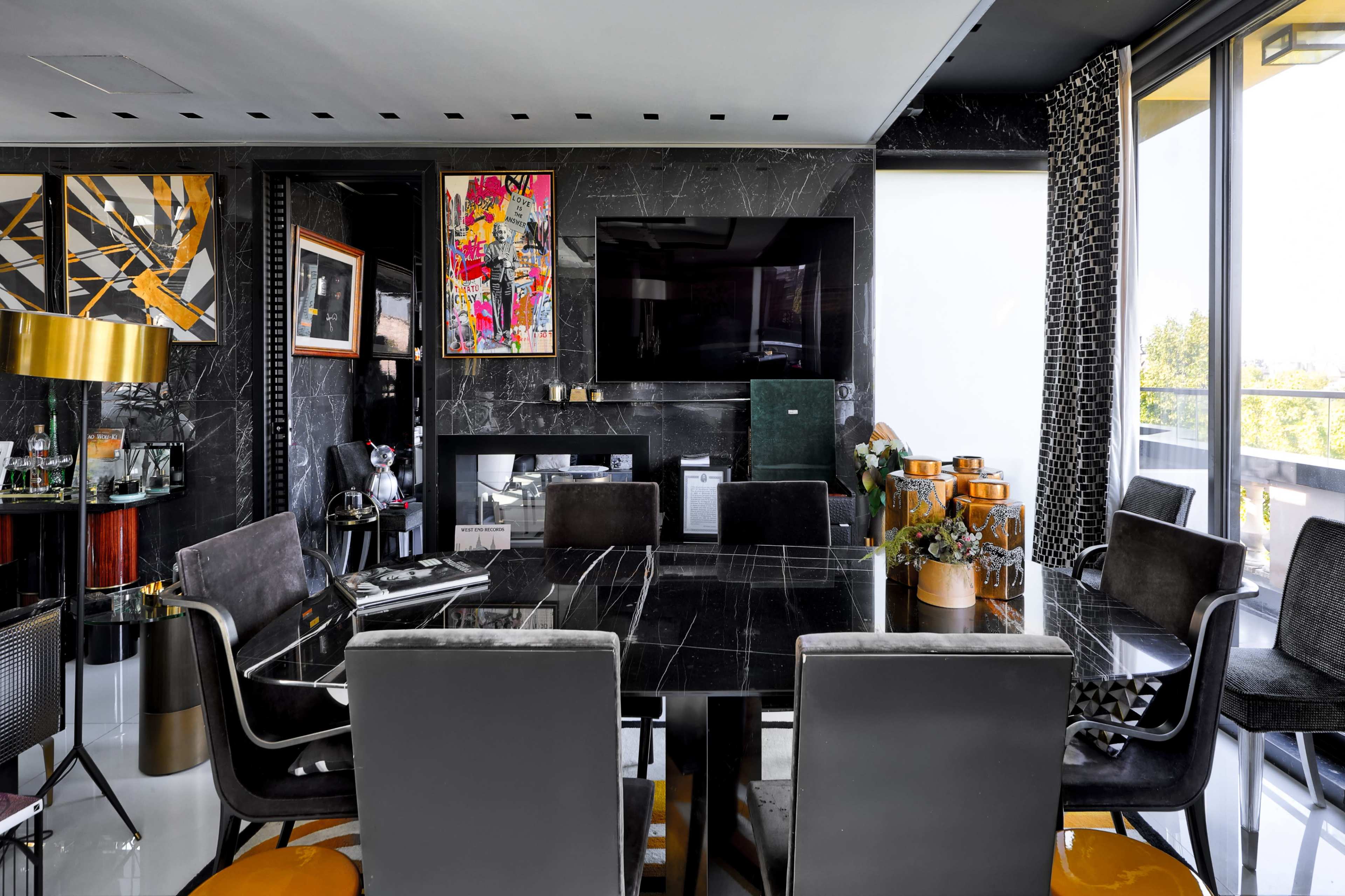 The image shows a modern dining area featuring a black marble table surrounded by dark chairs, with a large television mounted on the wall and colorful artwork displayed.