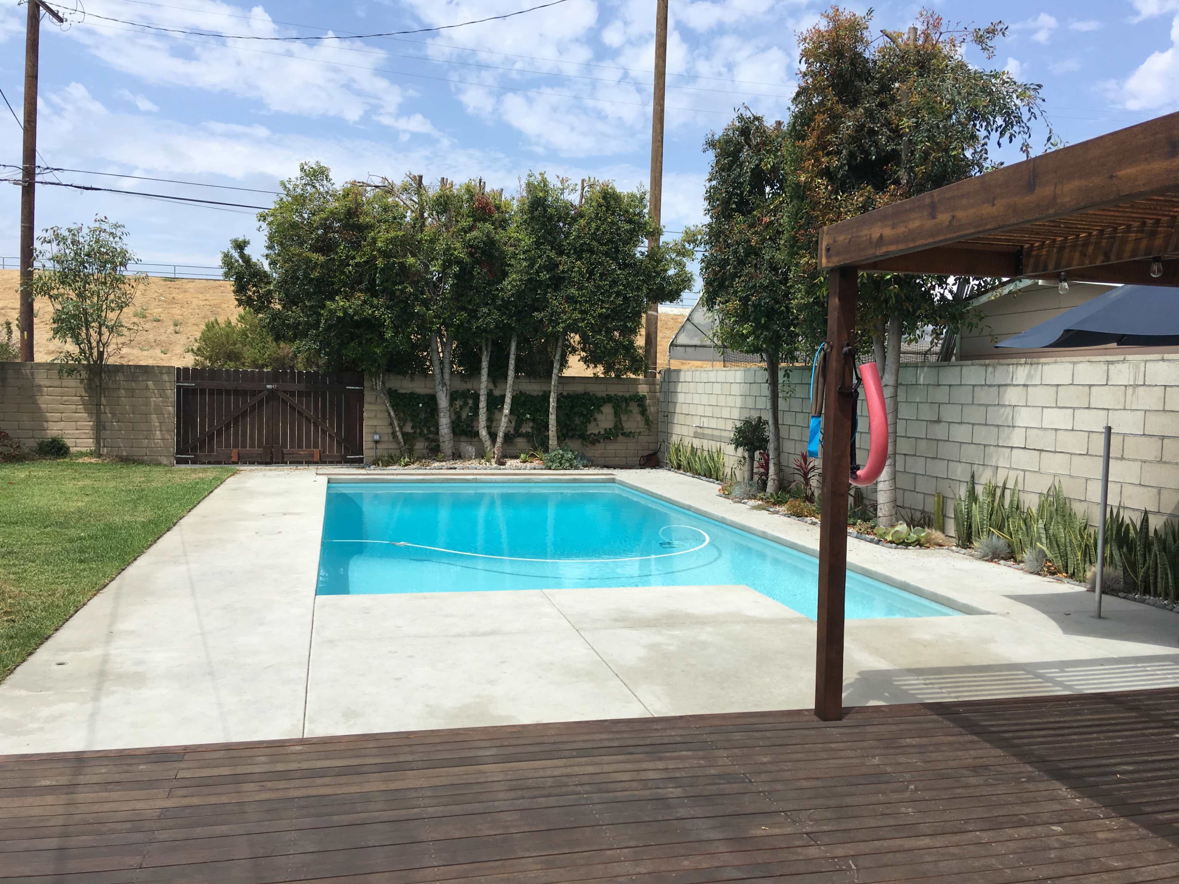 The image shows a backyard with a swimming pool surrounded by concrete and grass, with trees and a wooden pergola nearby.