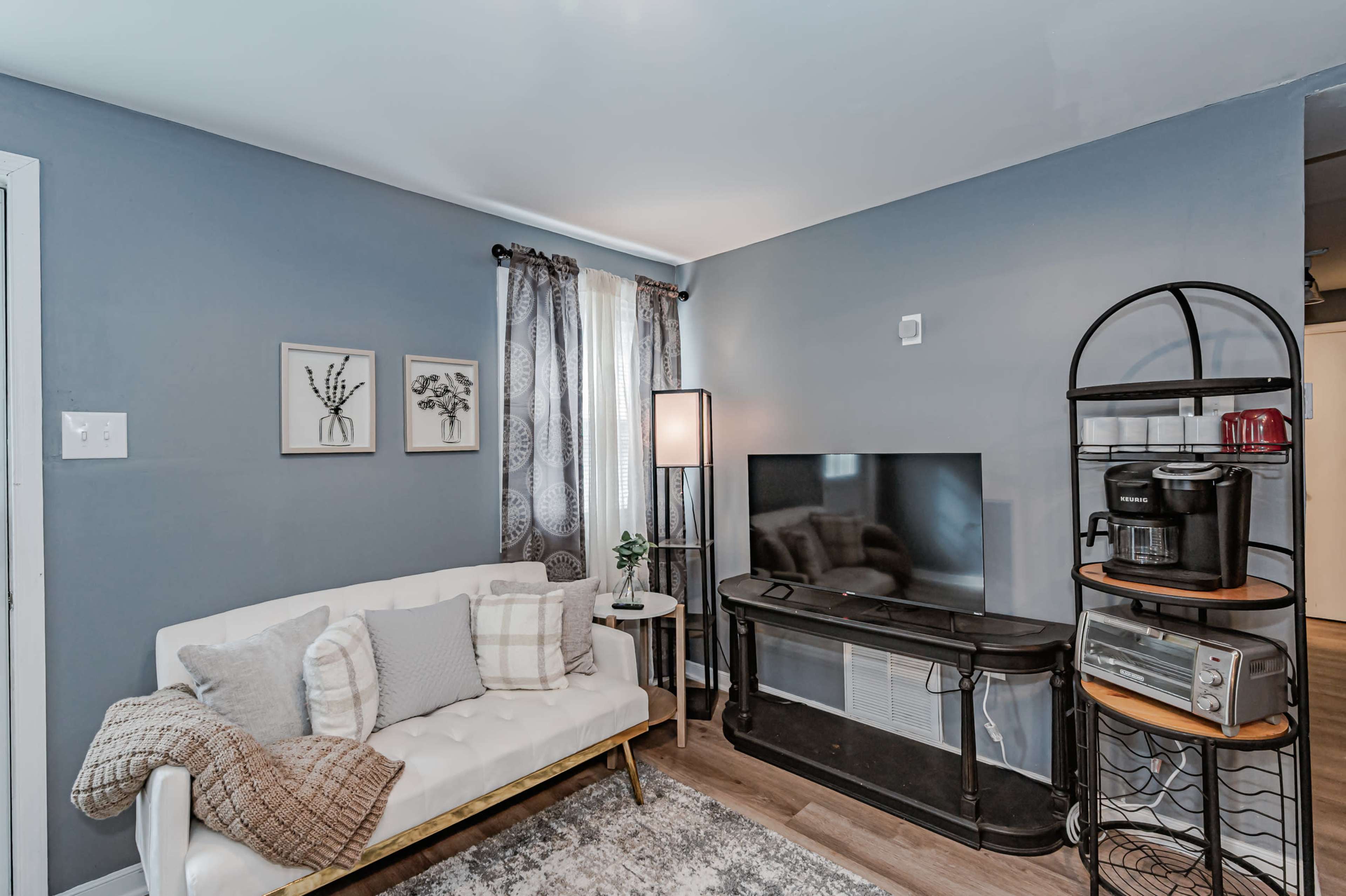 A small living room with a white couch, a television stand, and a coffee machine on a black shelf against a blue wall.