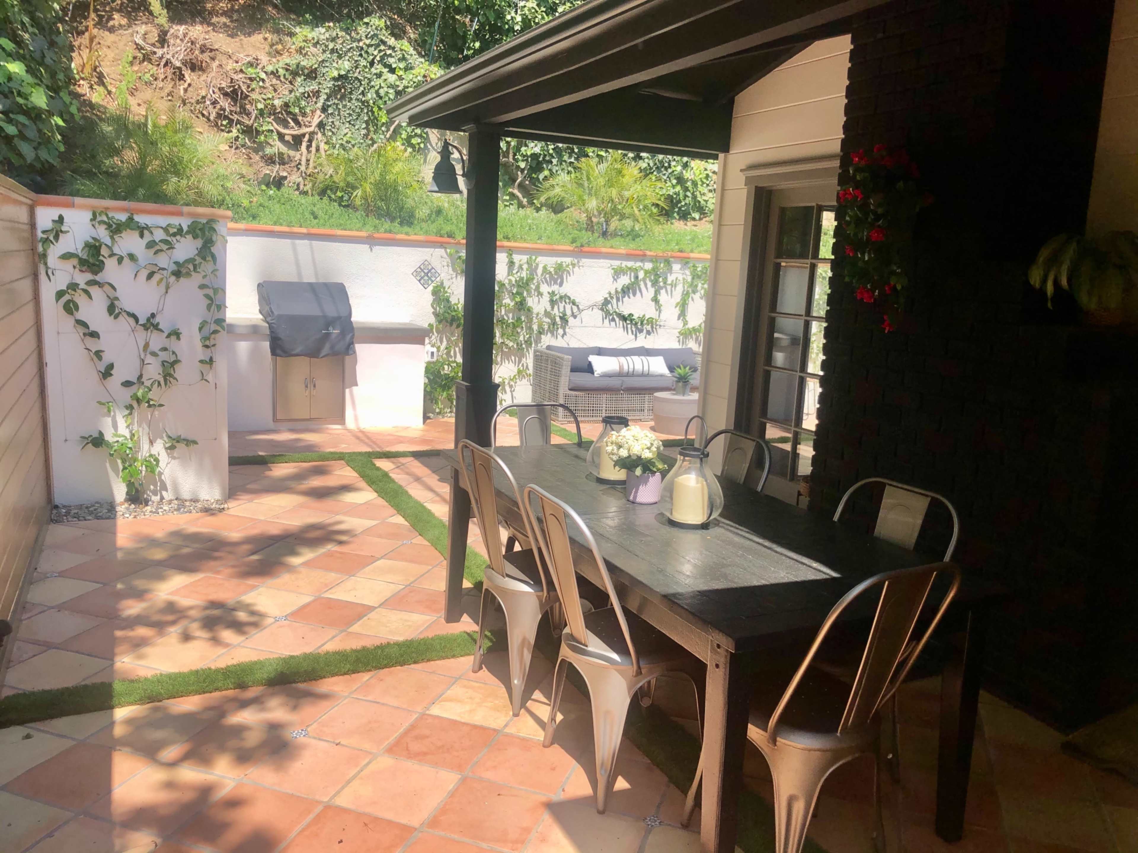 The image shows a patio area with a wooden dining table, metal chairs, a grill, and a lounge area surrounded by greenery and tiled flooring.
