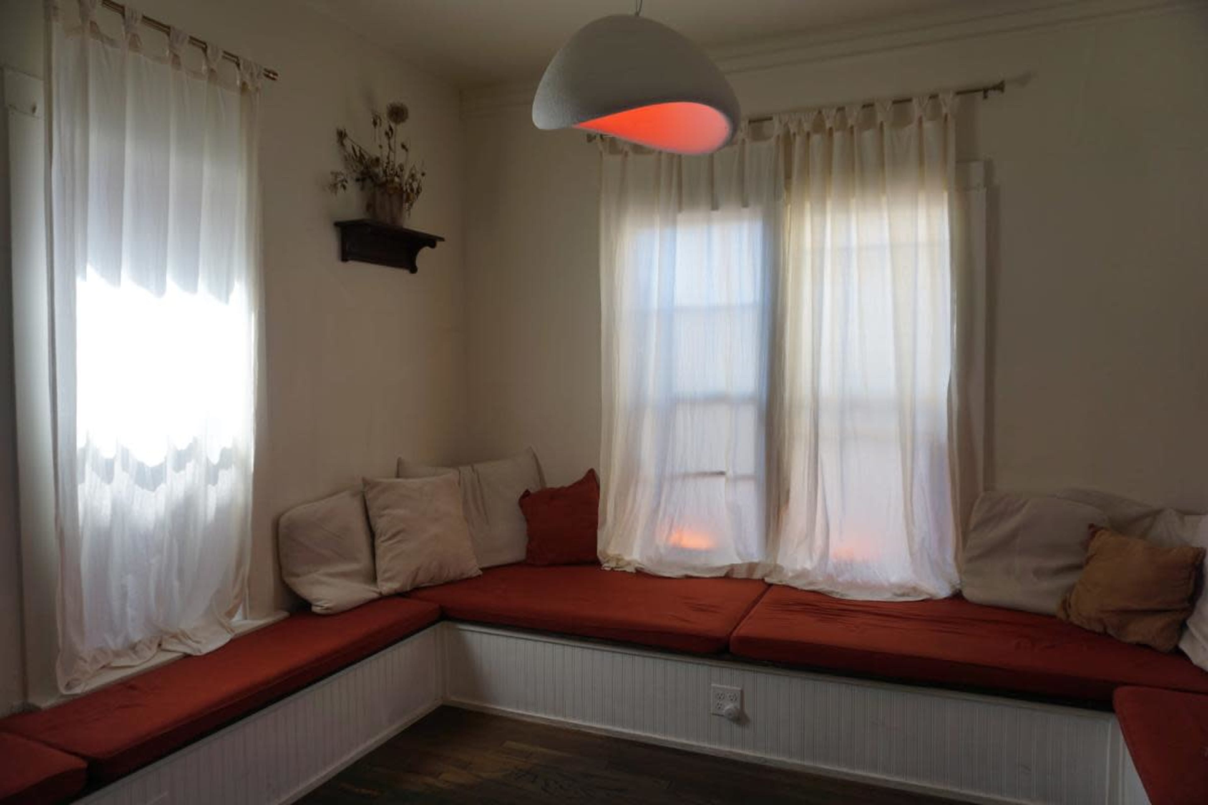 A cozy nook features a built-in bench with cushions, surrounded by two large windows adorned with sheer curtains.