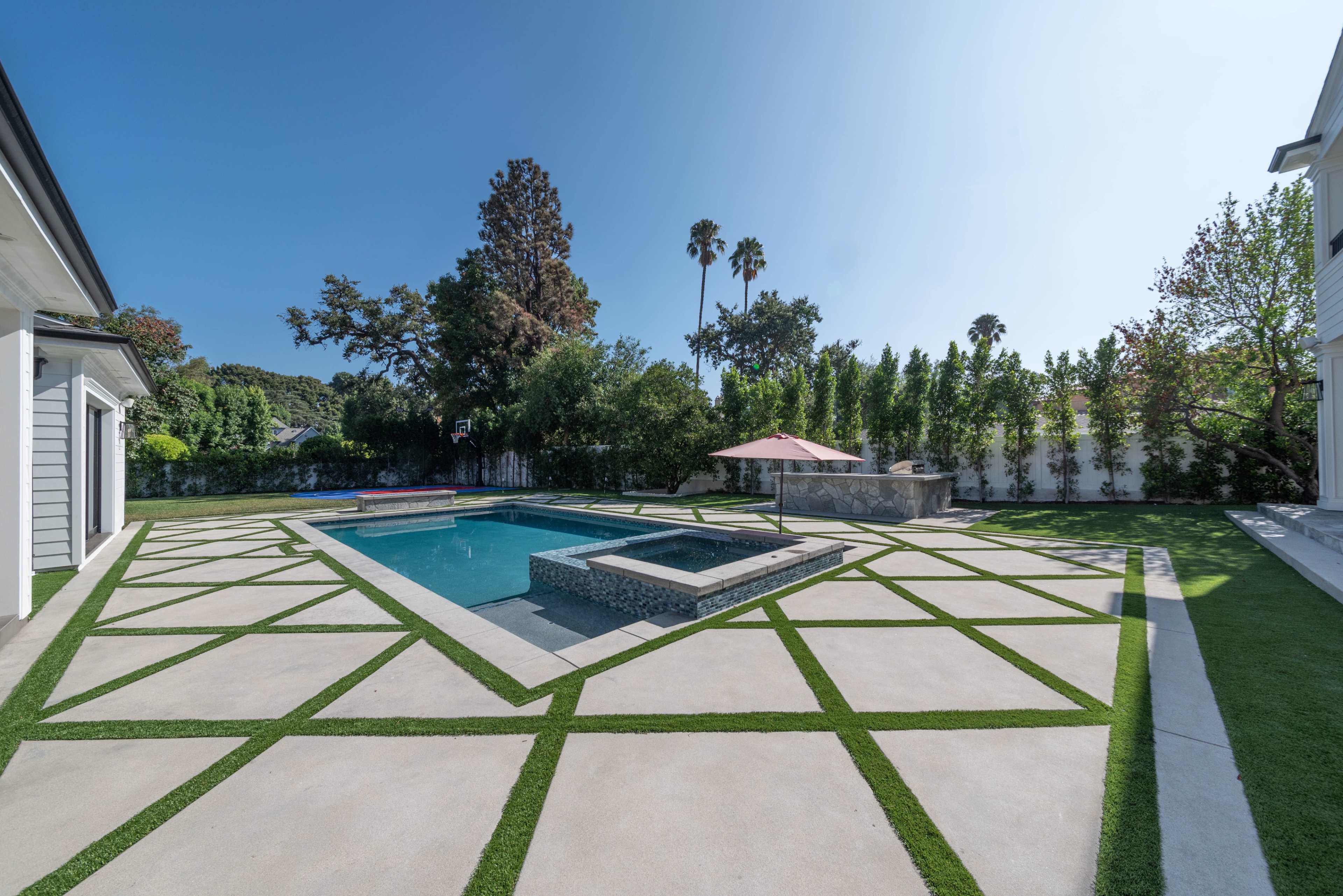 The image shows a geometric concrete patio with a swimming pool and a landscaped yard featuring trees and an umbrella.