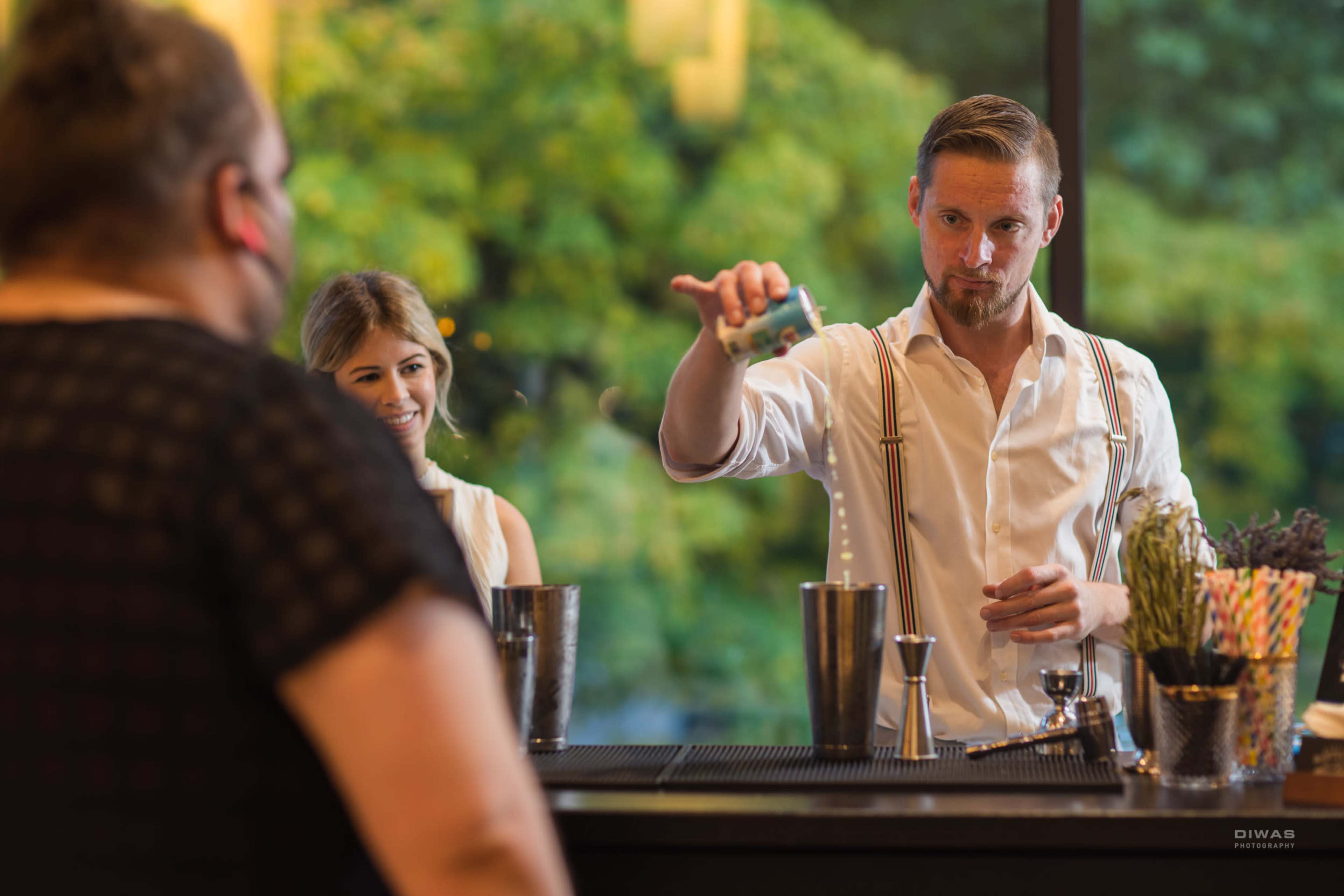 A bartender in a white shirt and suspenders is pouring a drink from a shaker while a woman watches from the other side of the bar.