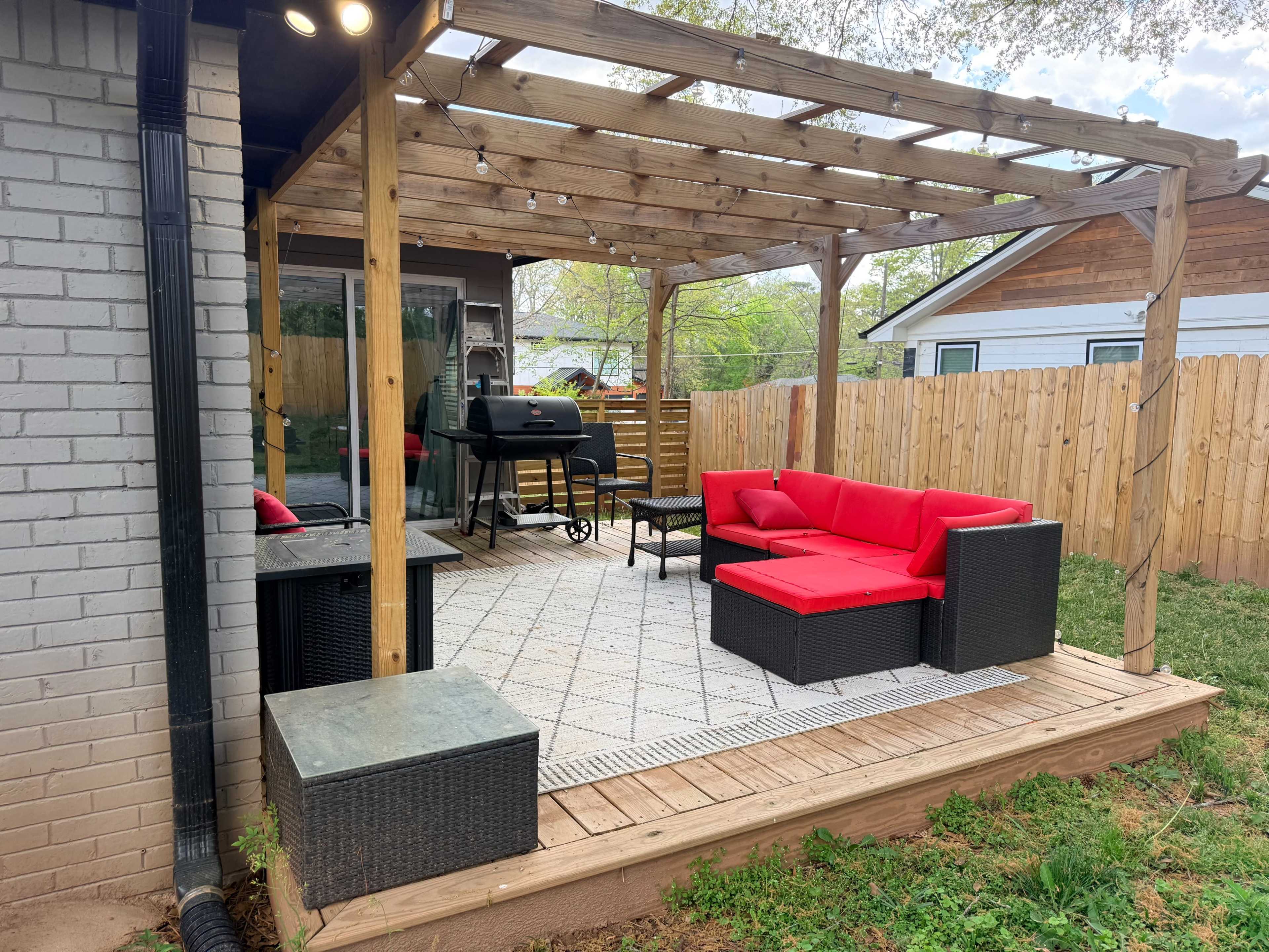 The image shows a patio with a wooden pergola, featuring a red couch set, two black lounge chairs, and a barbecue grill, surrounded by a wooden fence and grass.