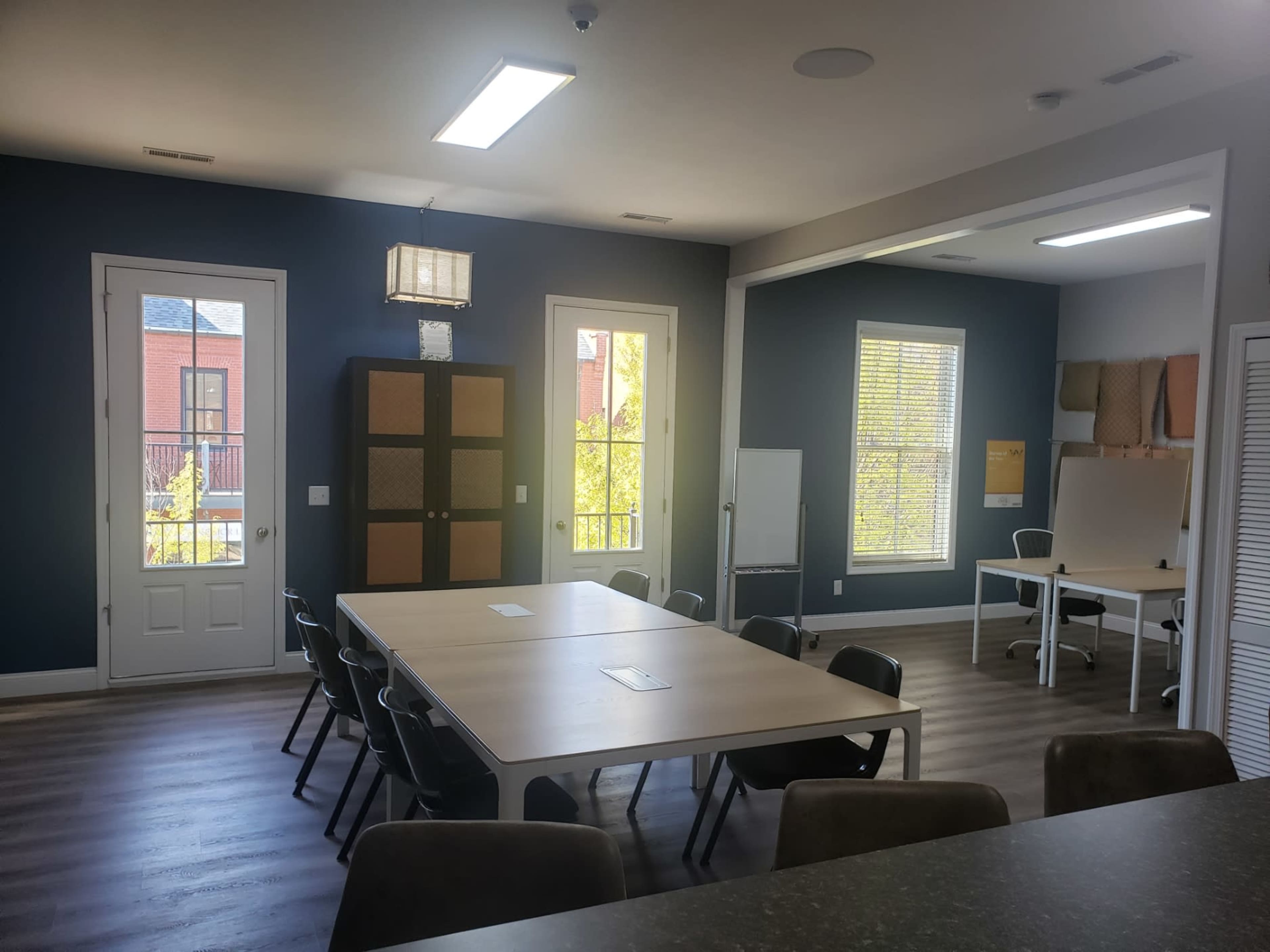 A brightly lit meeting room with a large table surrounded by chairs, open doors leading to an outdoor area, and a separate workspace visible in the background.