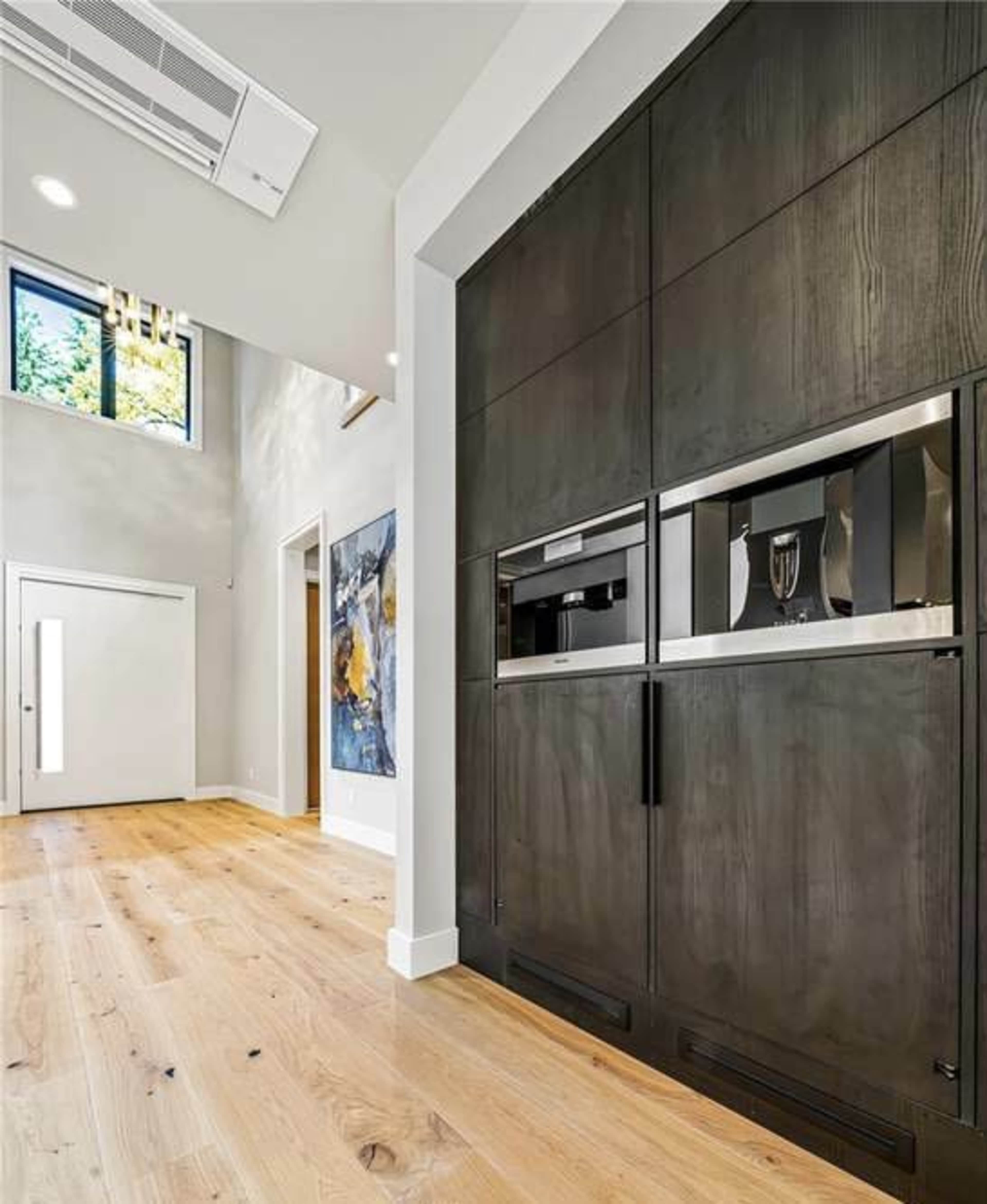 A modern interior hallway with a sleek, dark cabinetry wall and an entrance door, featuring a large window above and hardwood flooring.