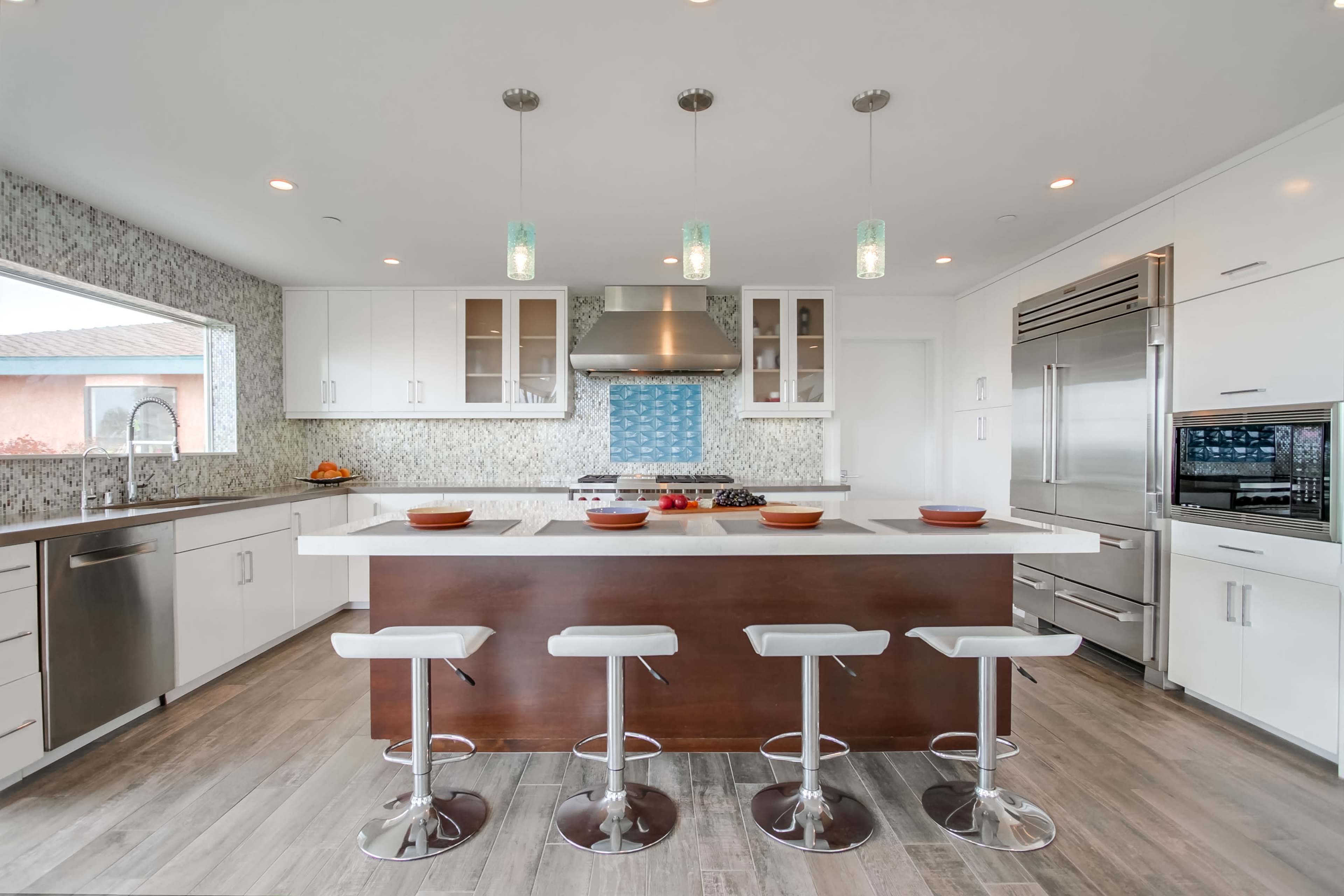 A modern kitchen featuring a large island with four white stools, stainless steel appliances, and light-colored cabinetry against a patterned backsplash.