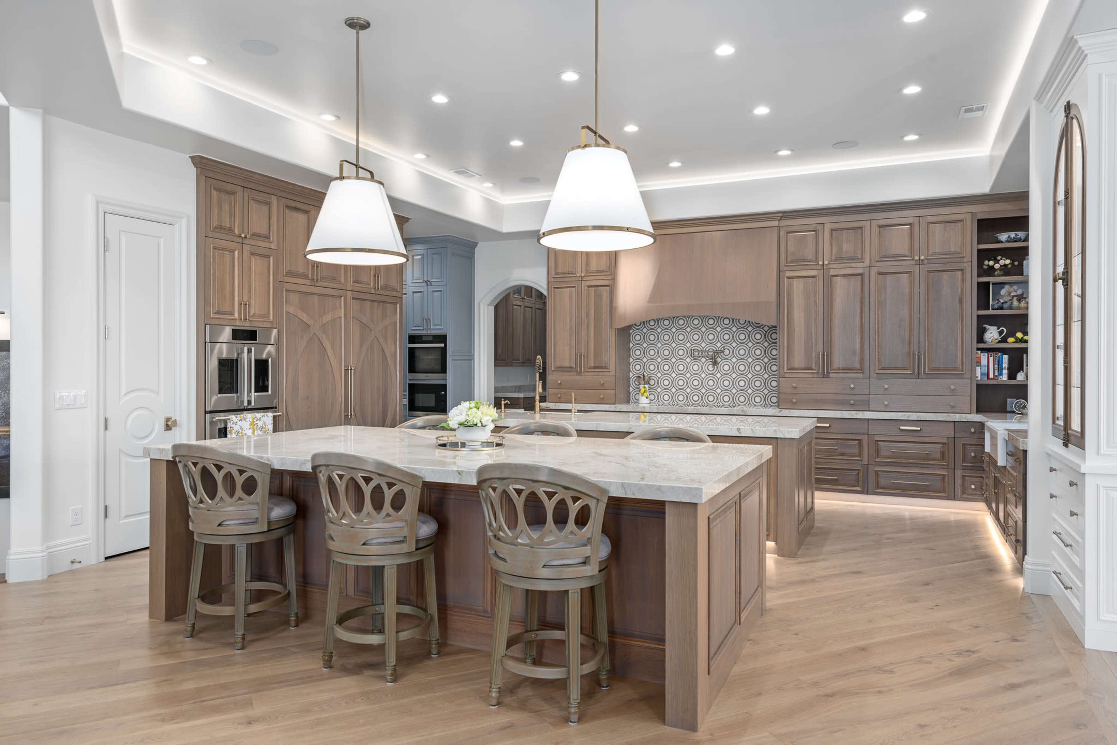 The image shows a modern kitchen featuring a large central island with four wooden bar stools, light fixtures hanging above, and cabinetry in various shades of brown.