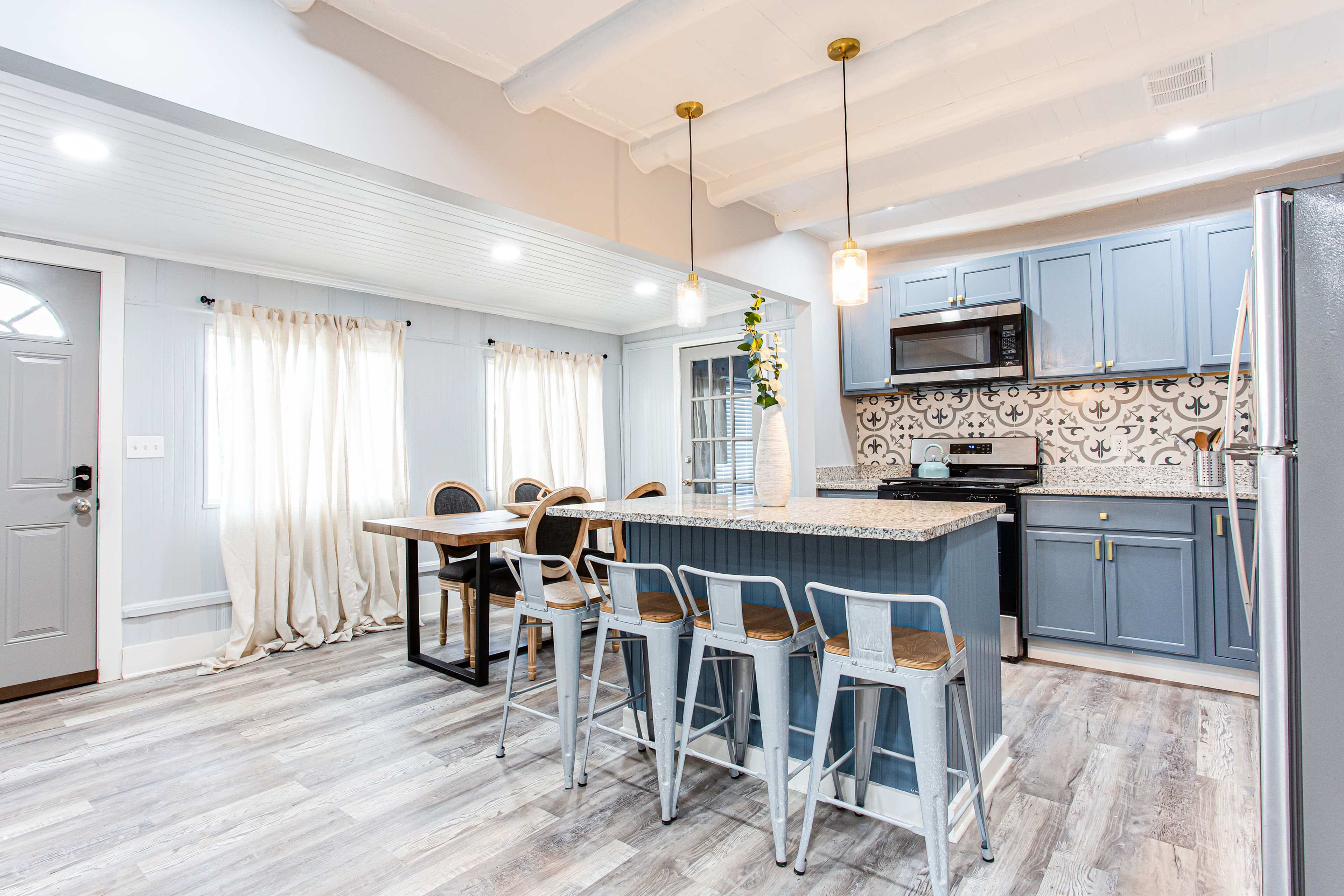 A modern kitchen features blue cabinetry, a large island with bar stools, and a dining area with a wooden table next to large windows.