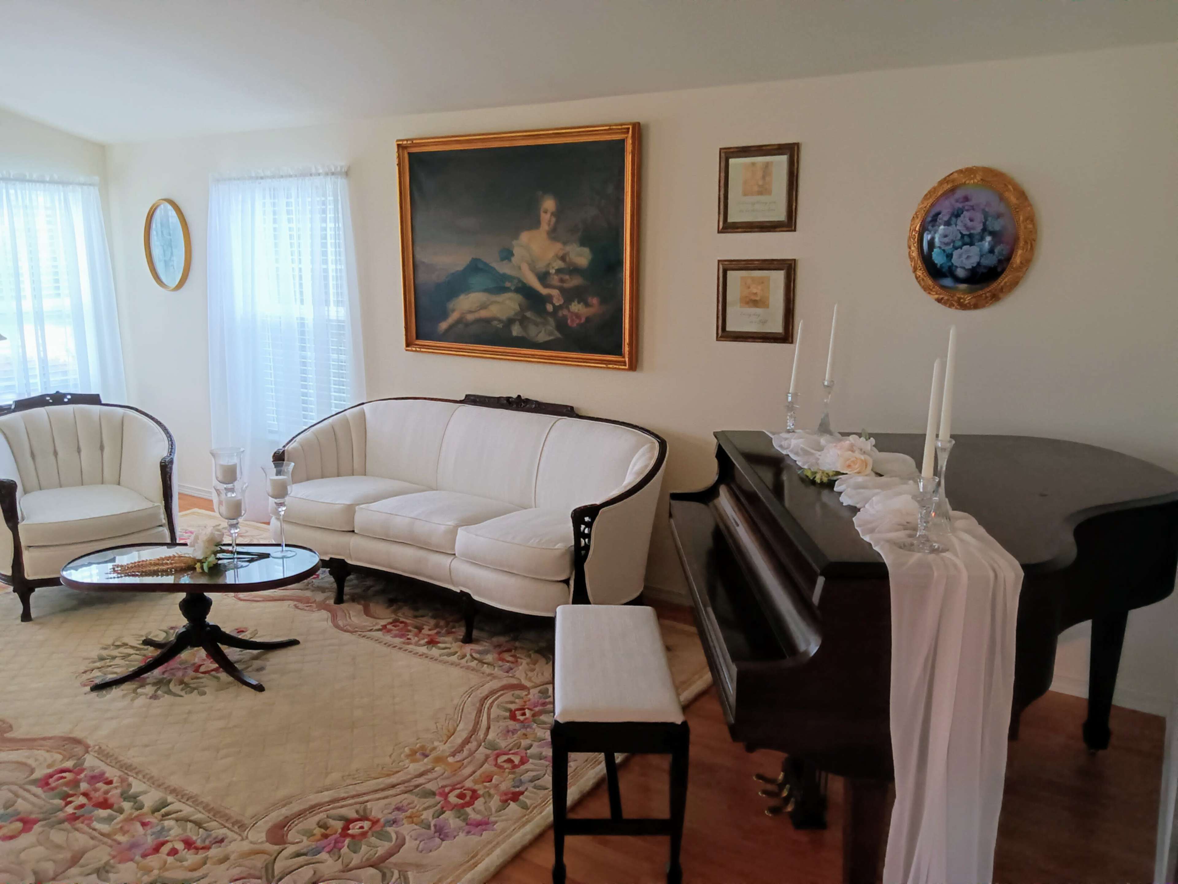 The image shows a bright living room featuring a vintage white sofa set, an ornate piano, and decorative artwork on the walls.