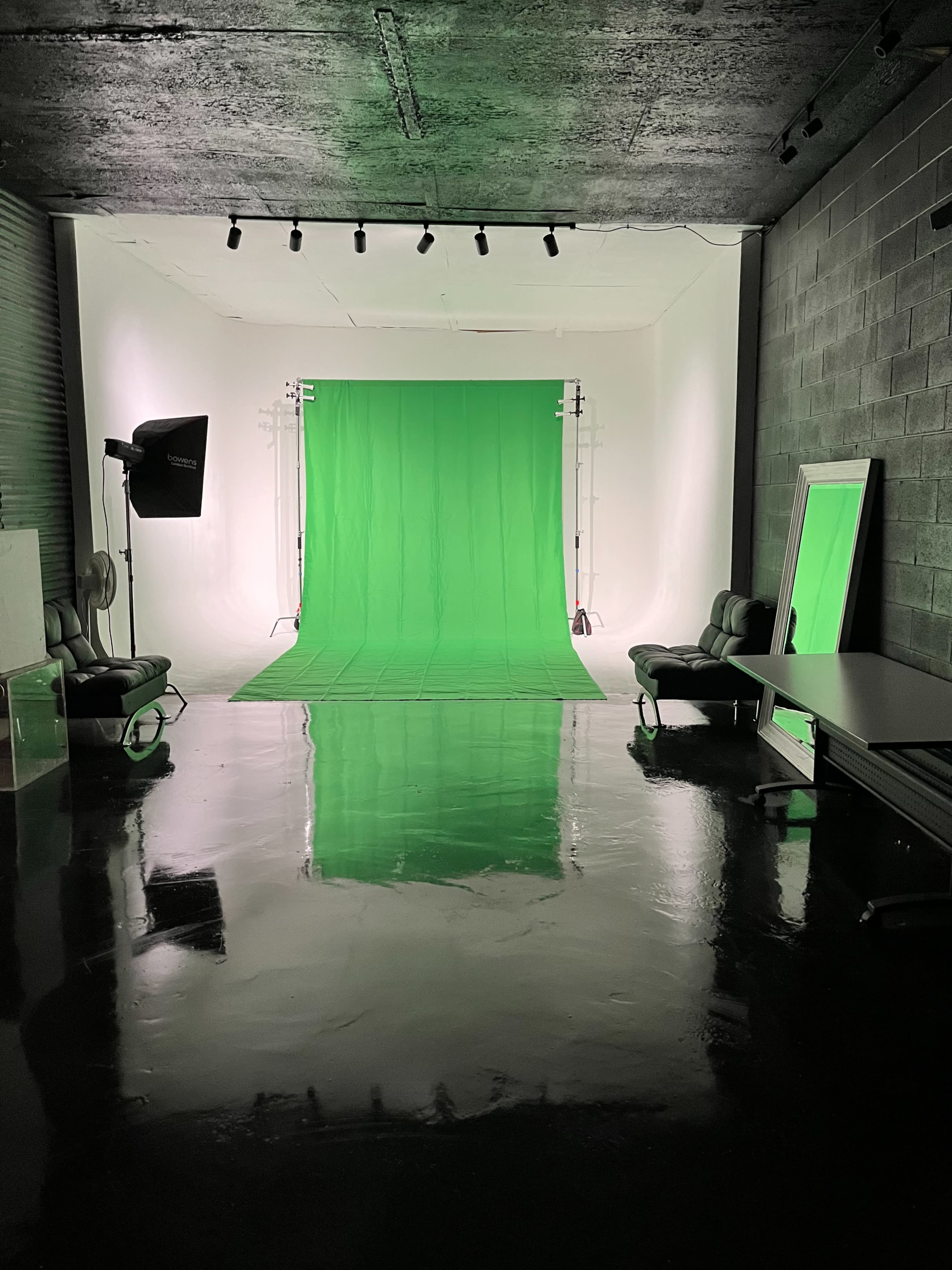 A photography studio with a green backdrop, equipped with lights, chairs, and reflective surfaces on the floor.
