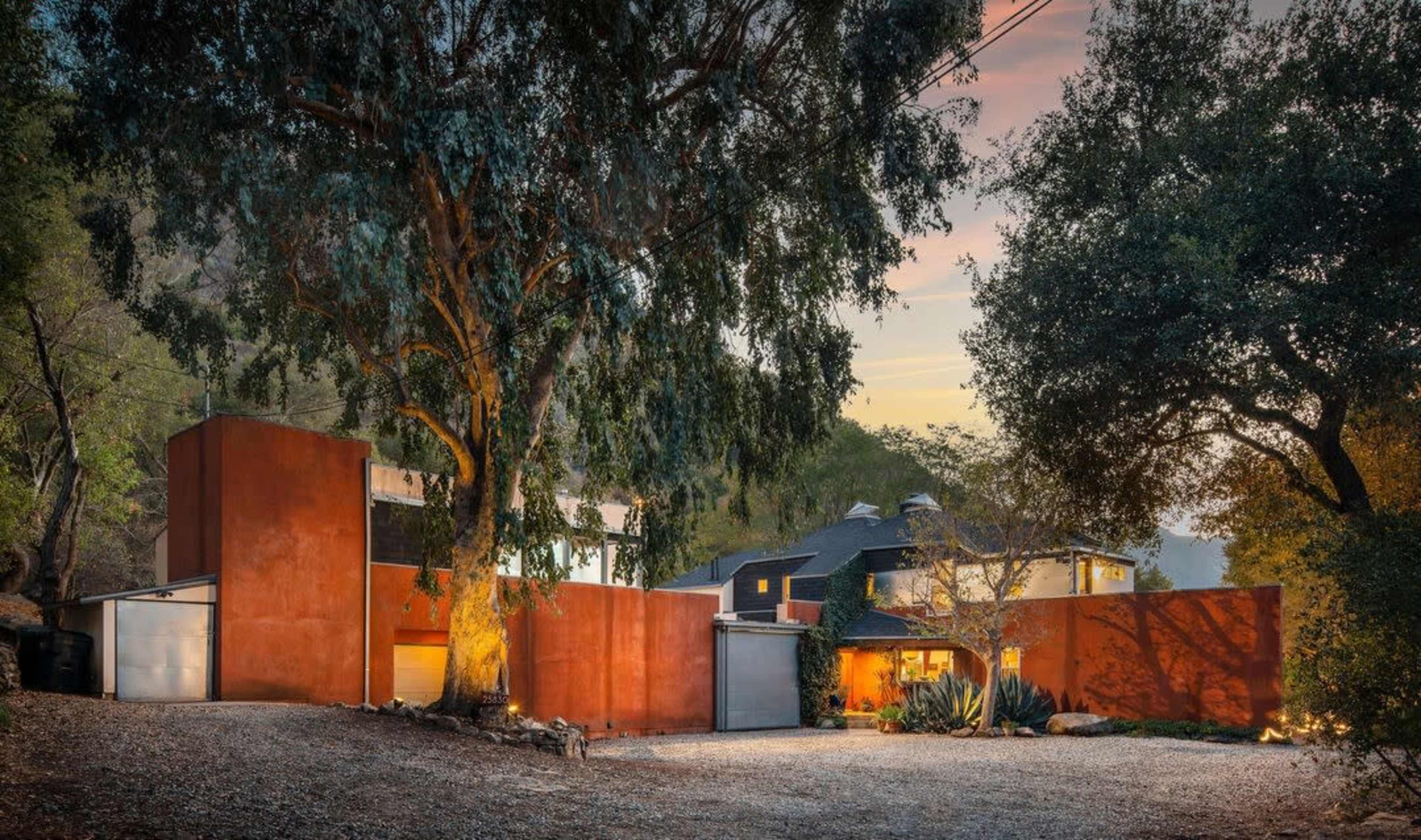A modern house with a rusty-colored exterior is surrounded by trees and gravel, set against a twilight sky.