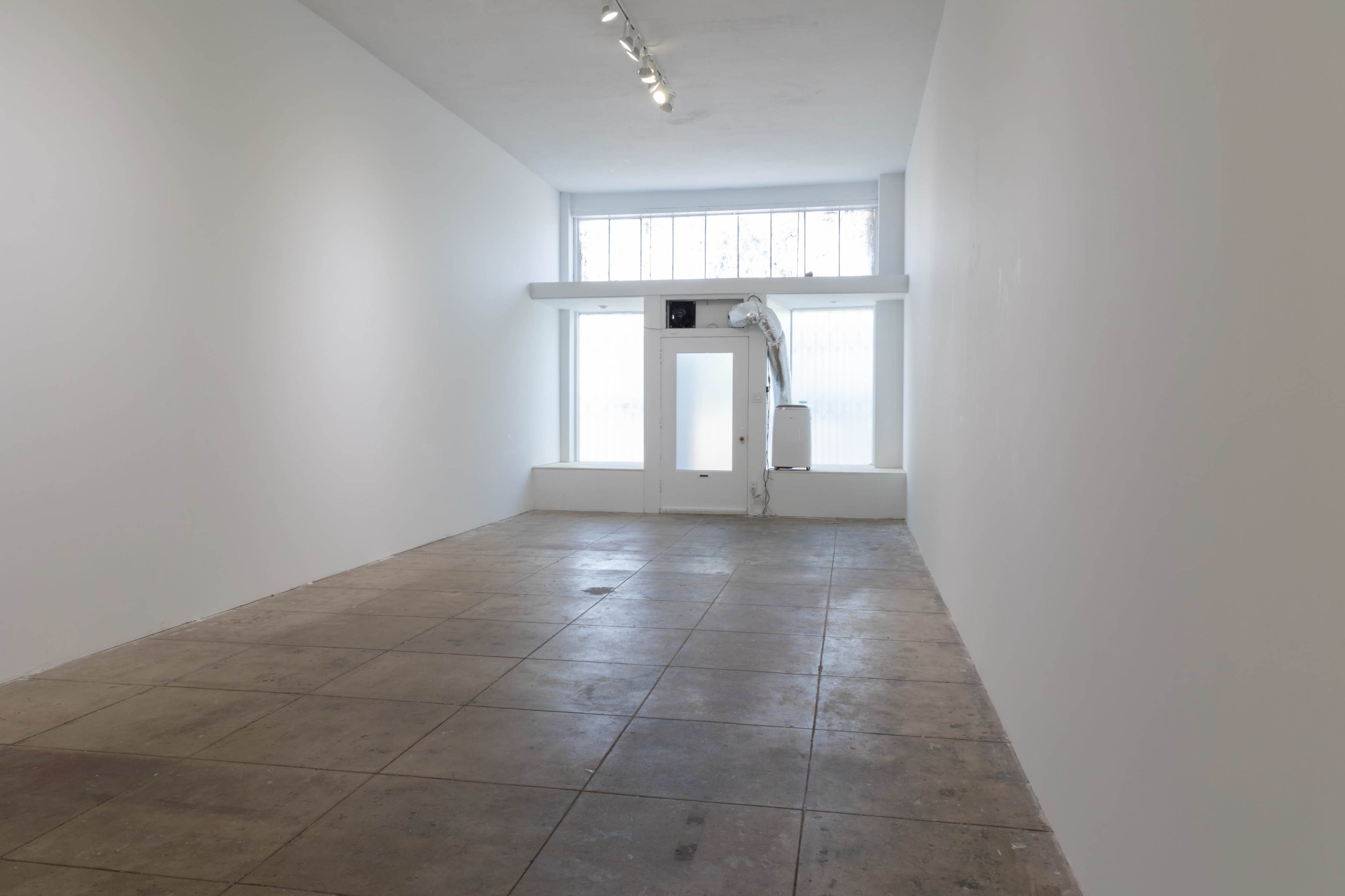 A sparse, empty interior space with white walls, large windows, and a concrete floor.