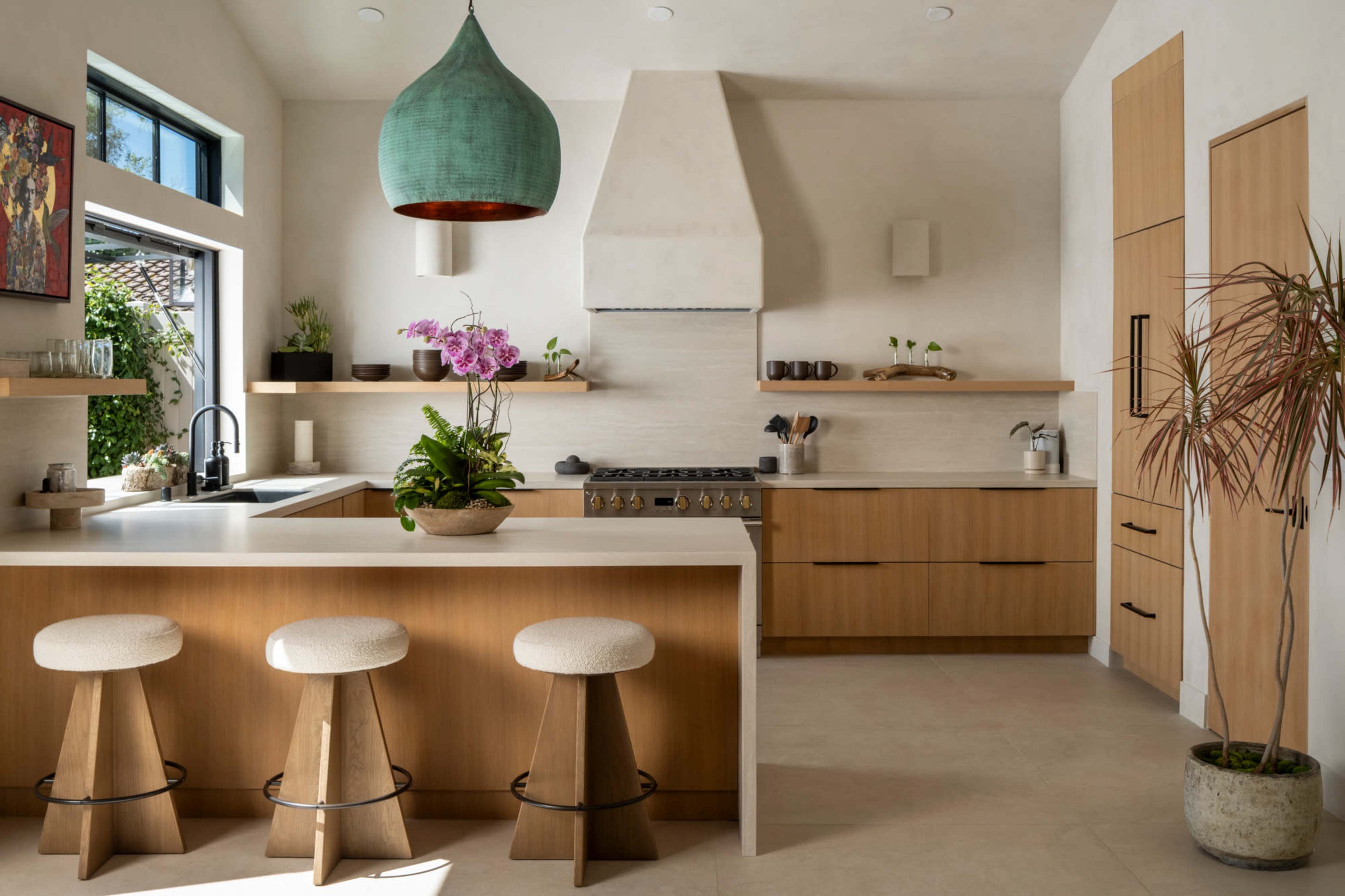 A modern kitchen features wooden cabinetry, a large island with three stools, and a pendant light above, accented by a countertop filled with plants and kitchenware.