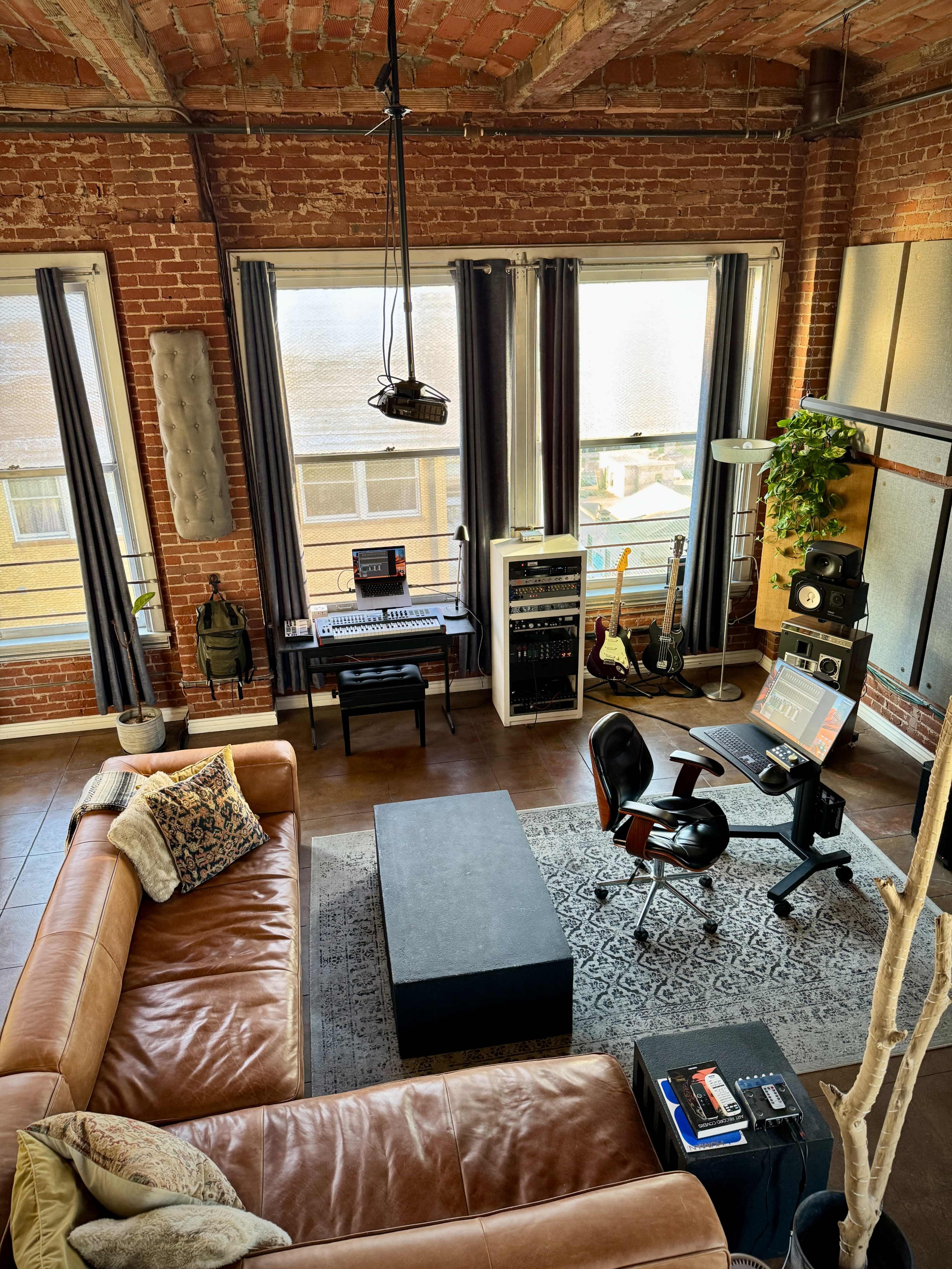 The image shows a modern living space with exposed brick walls, large windows, a brown leather sofa, a black coffee table, an office setup with a computer, and musical instruments scattered around.