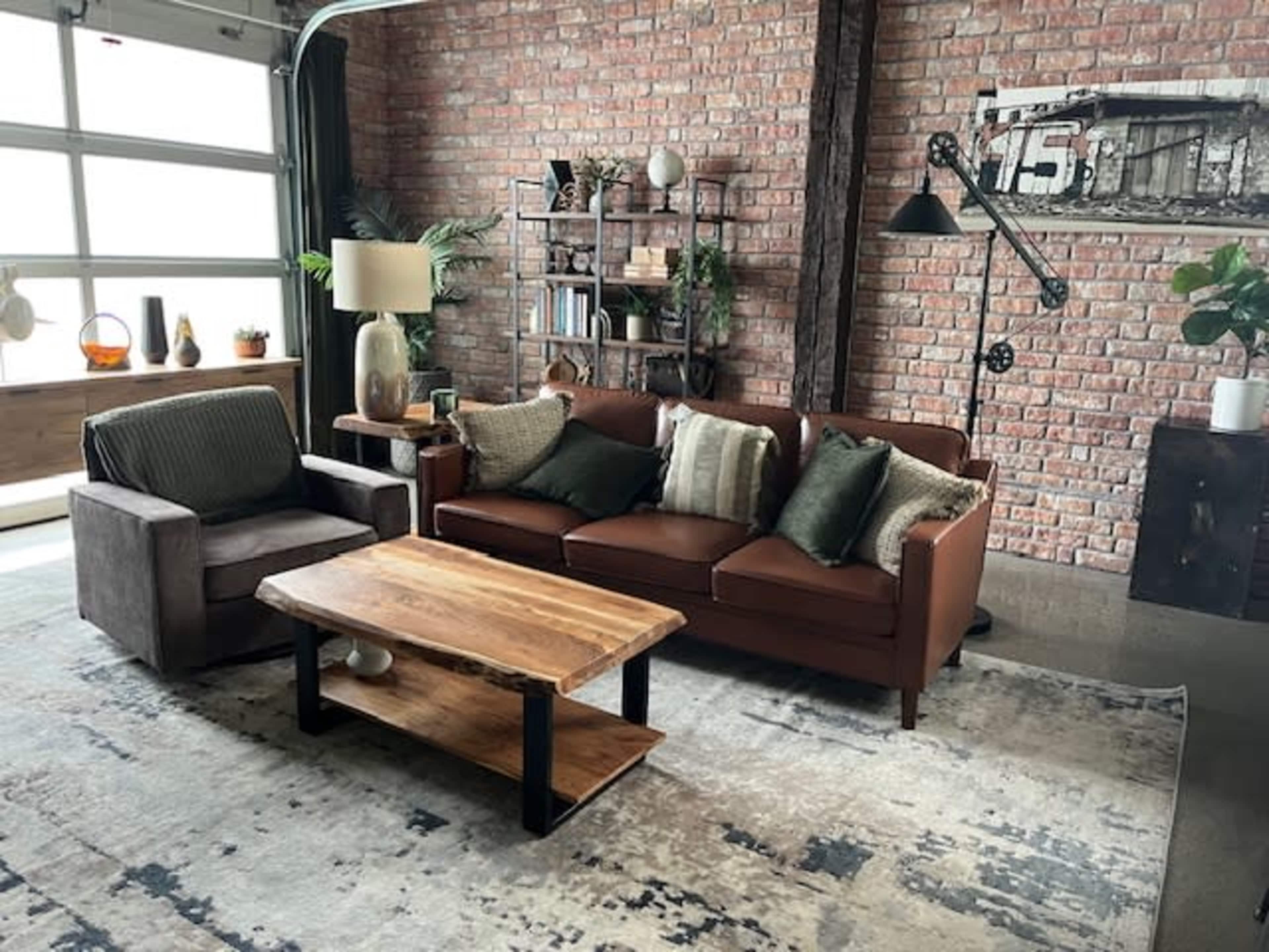 The image shows a modern living room with a brown leather sofa, a gray armchair, a wooden coffee table, and brick walls adorned with decorative plants and shelves.