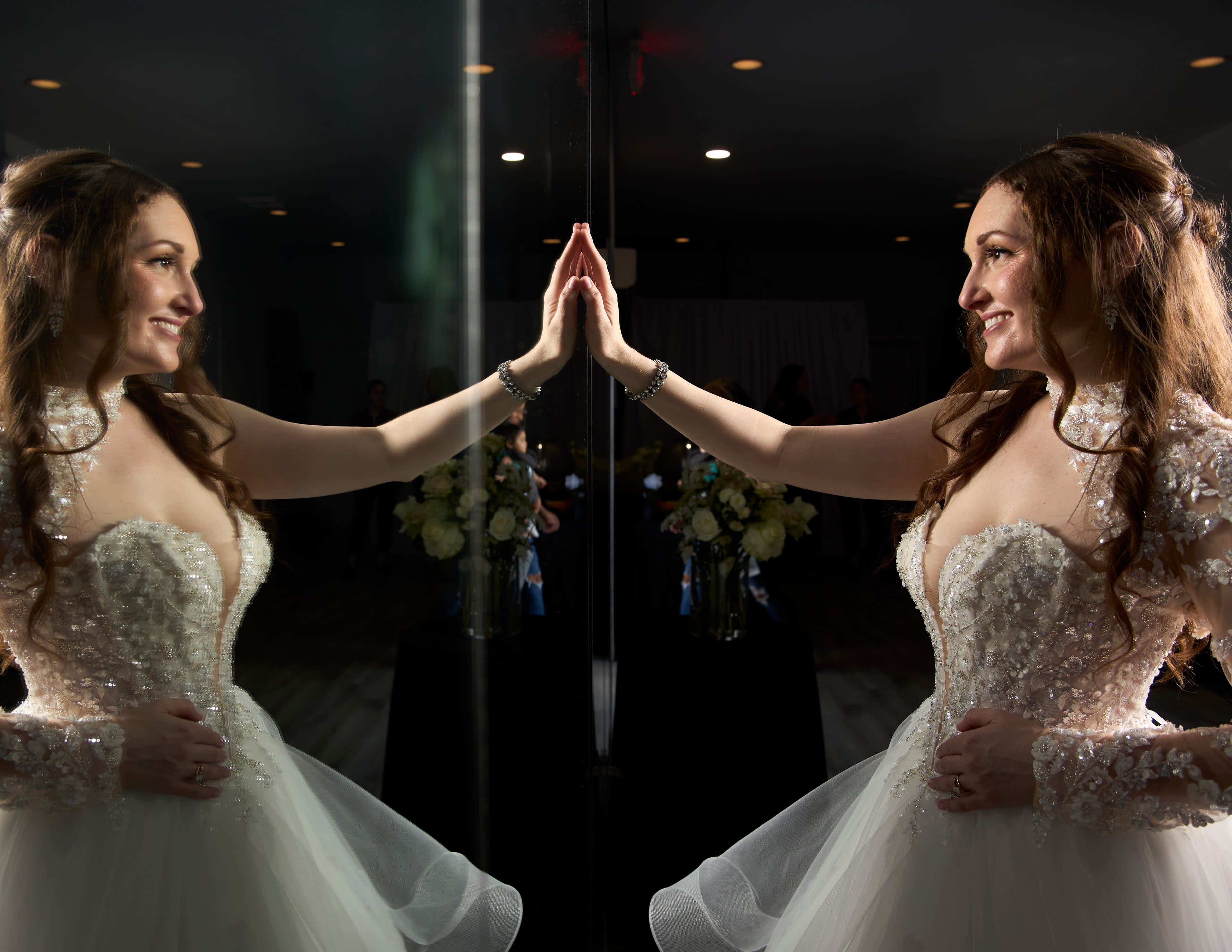 A young woman in a white wedding dress poses while touching a mirror, reflecting her image.