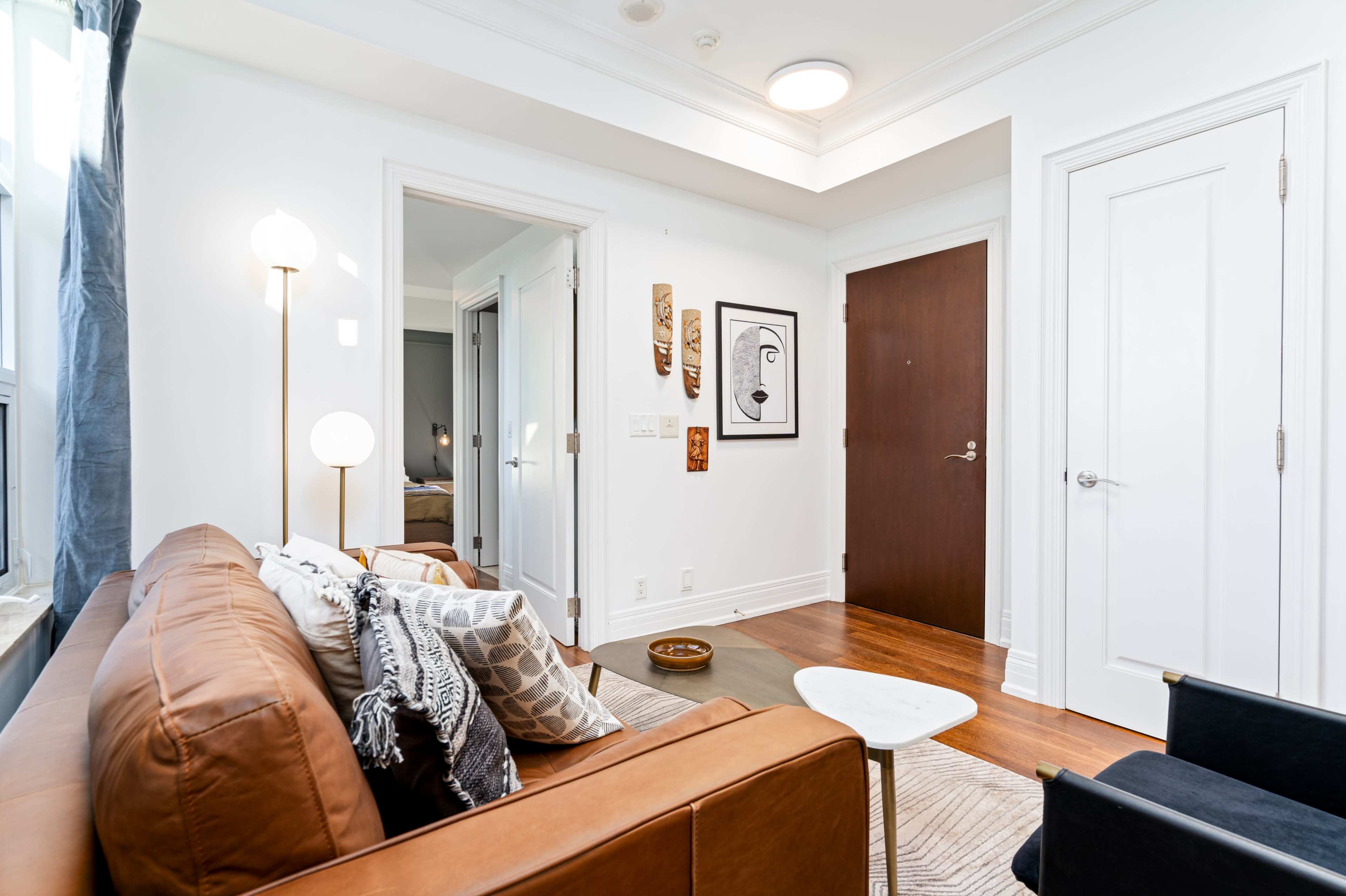 A modern living room with a brown leather sofa, a small white coffee table, and a framed artwork on the wall, leading to a closed door.